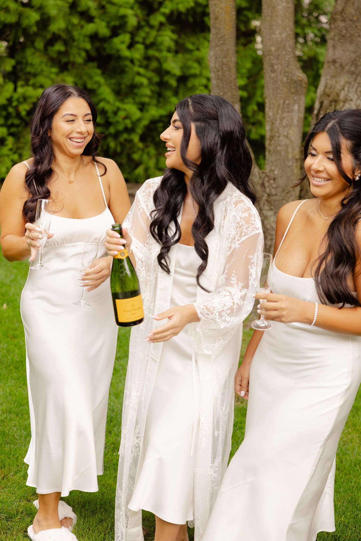 A group wearing white dresses laughs and celebrates while holding champagne glasses in a garden setting.