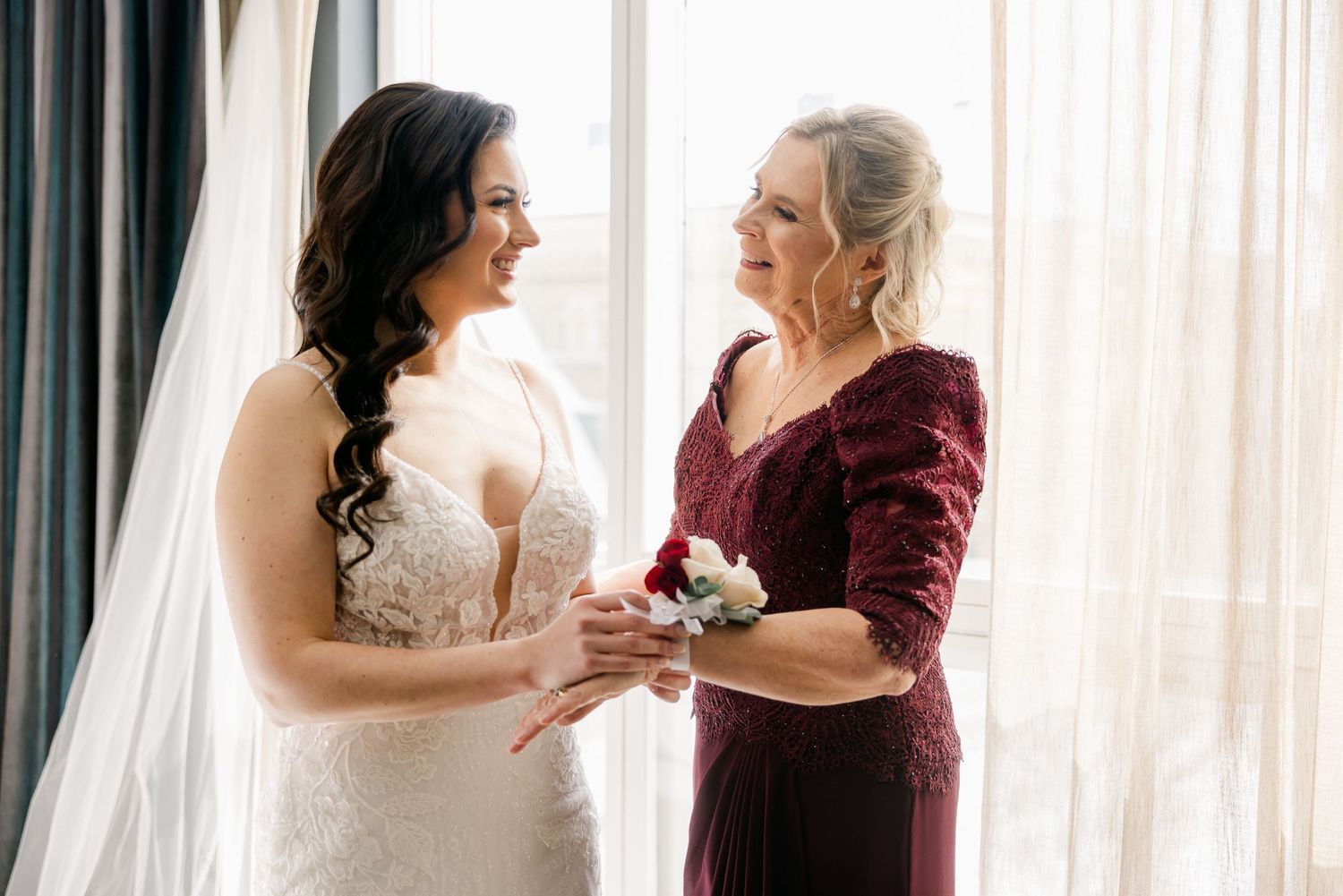 Candid moment of preparation between bride in white gown and helper in burgundy dress.