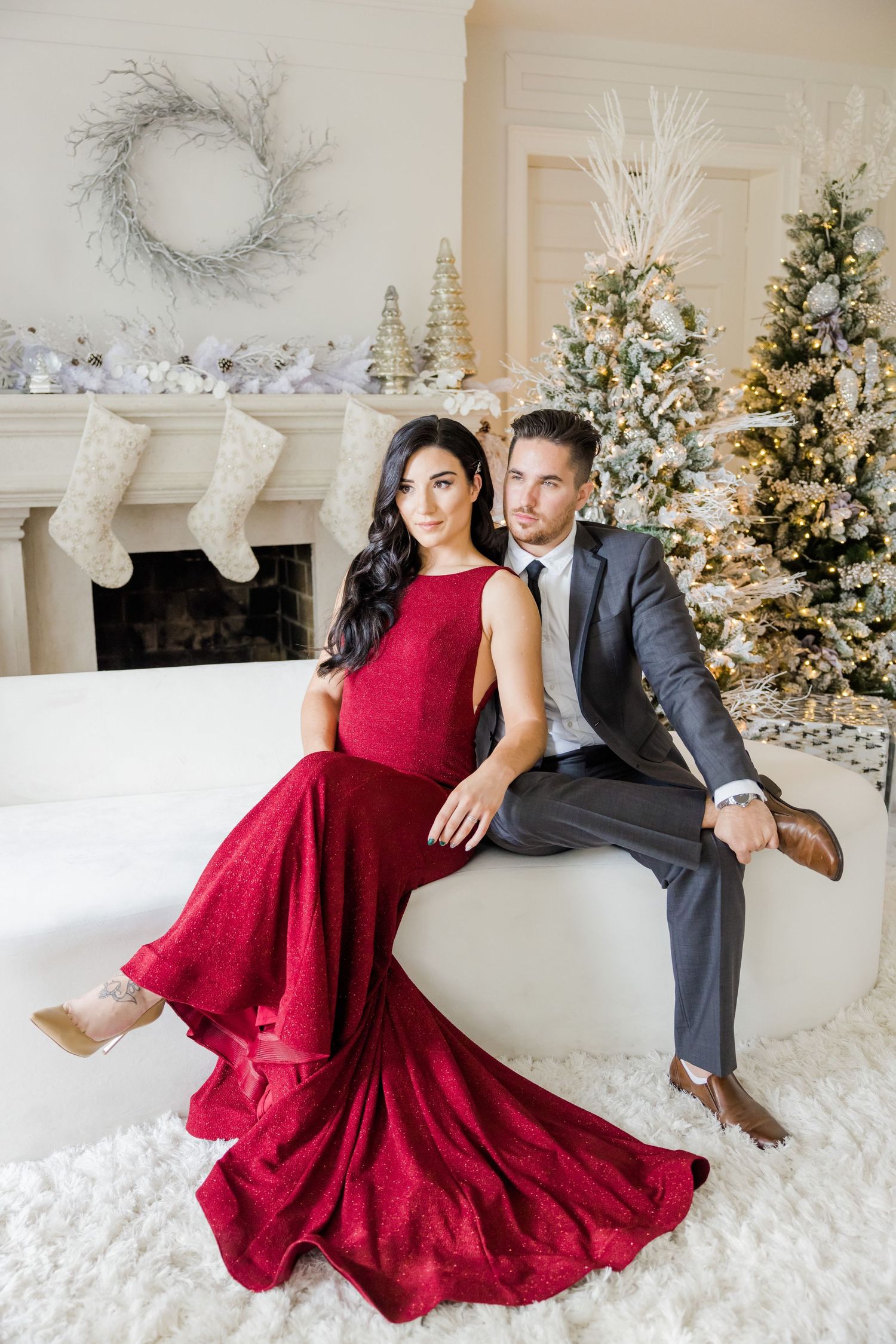 Glamorous holiday photoshoot with flowing red gown against white Christmas decor and trees.