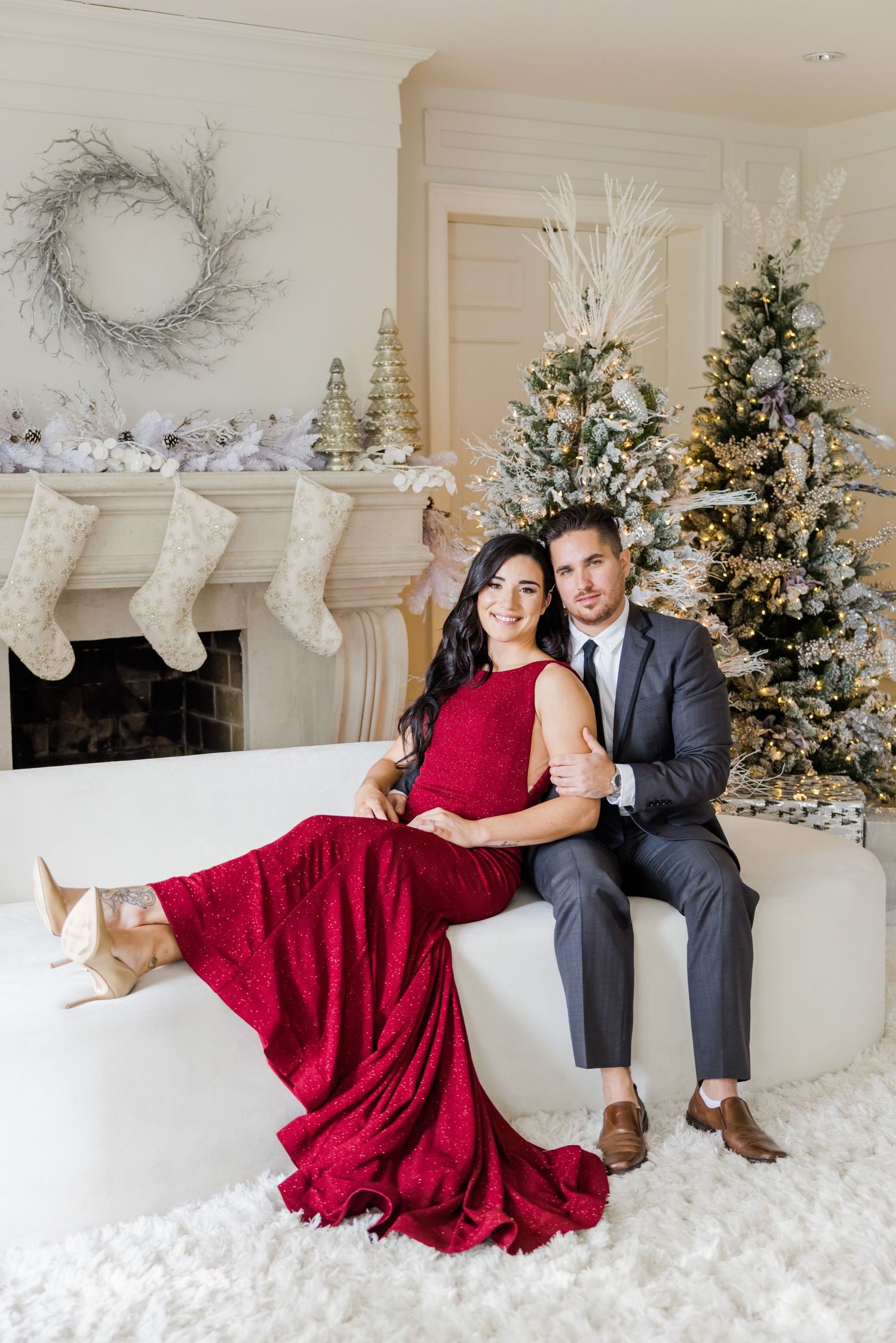 Intimate holiday portrait with flowing red dress against Christmas trees and decorated mantle.
