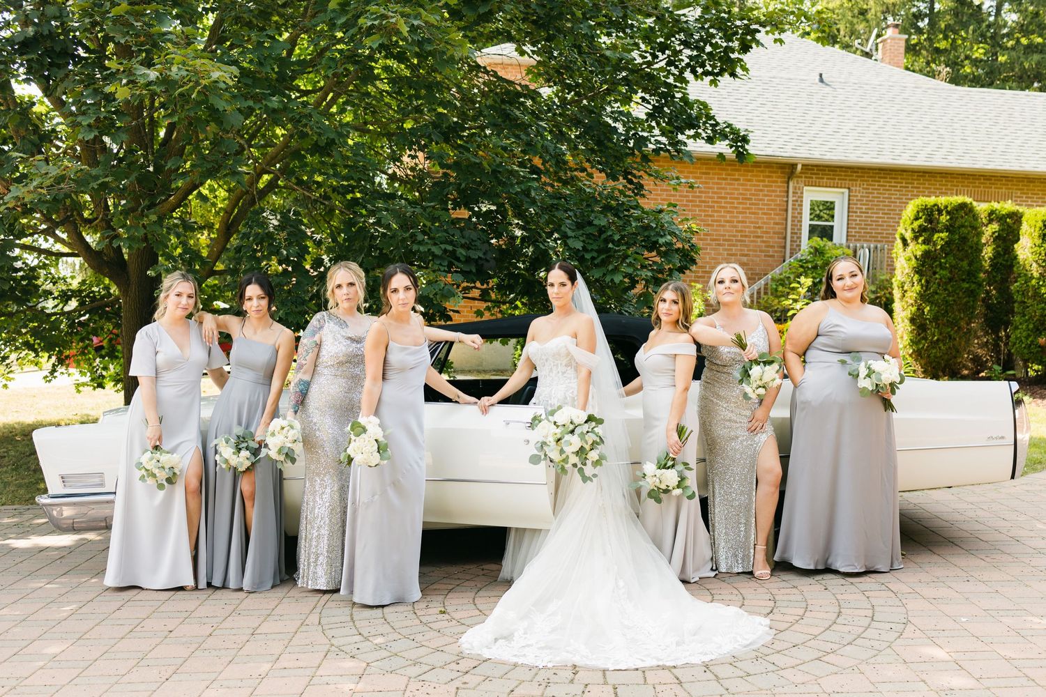 Bridal party in silver dresses posed with vintage white car against brick and garden backdrop.
