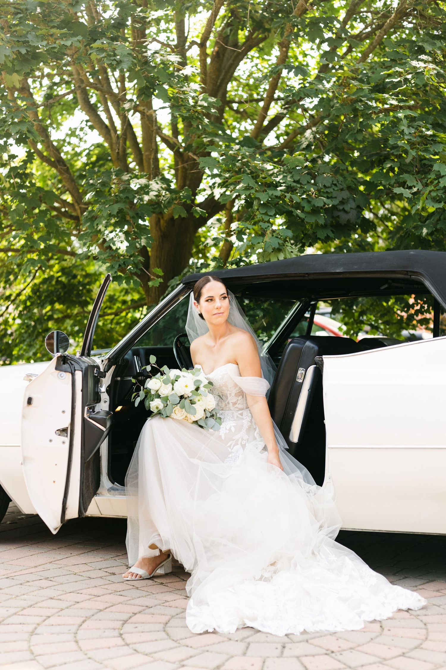 Classic white wedding dress and bouquet against vintage Cadillac and green trees.
