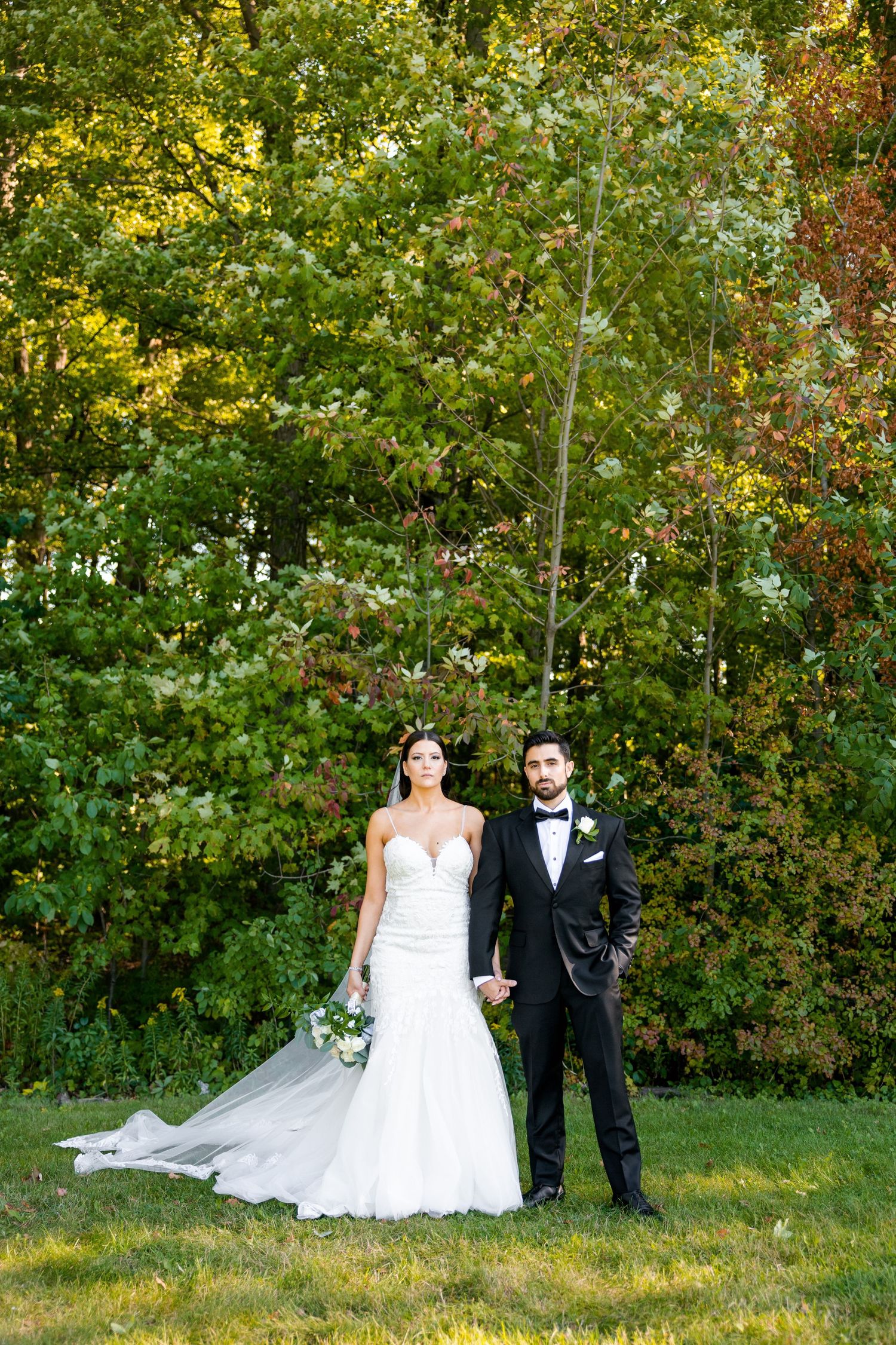 A formal wedding portrait of a couple standing in front of lush green trees on their special day.