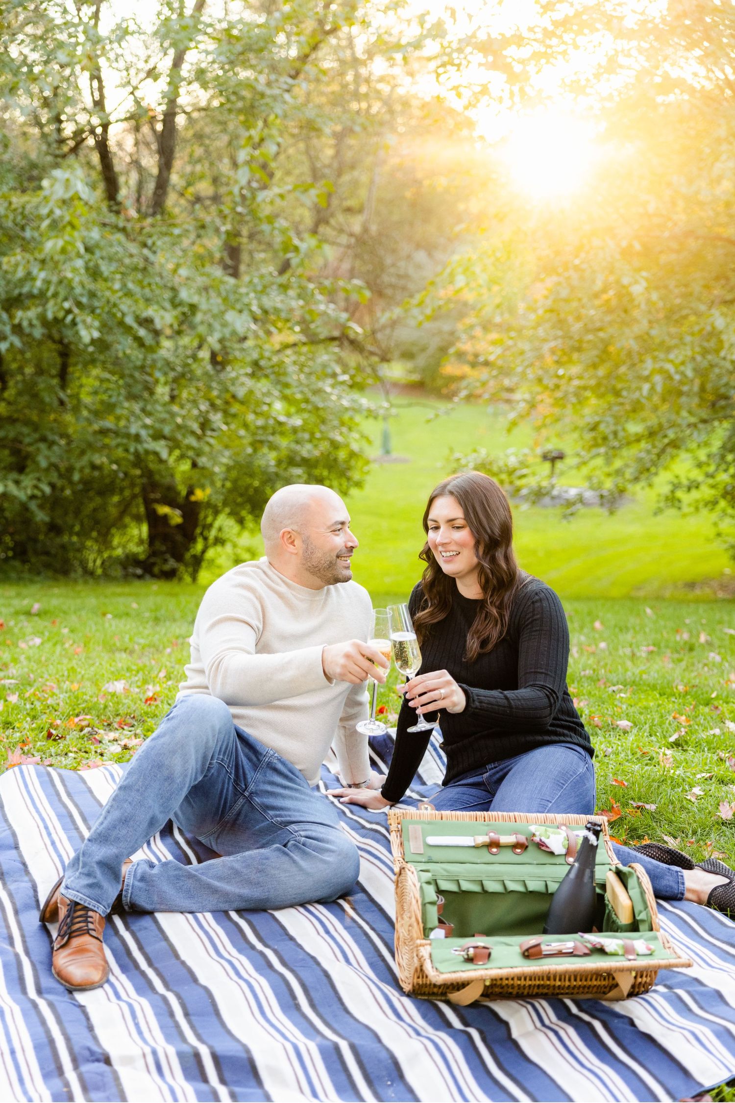 Couple shares drinks during picnic with vintage basket on striped blanket in sunlit park.