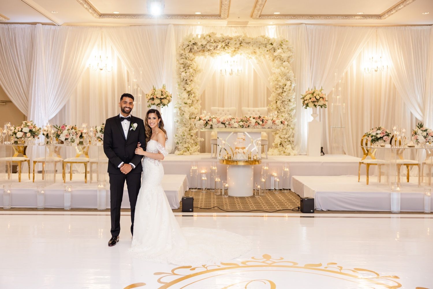 Wedding ceremony setup with white floral arch and gold chairs in an elegant ballroom.