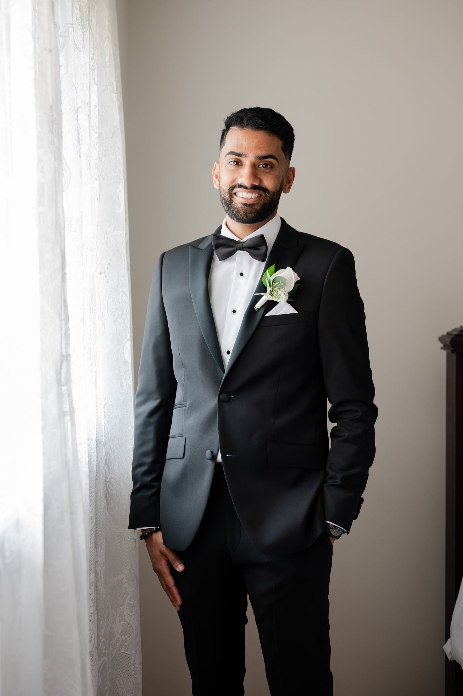Elegant groom in classic black tuxedo with white boutonniere smiles for formal portrait.