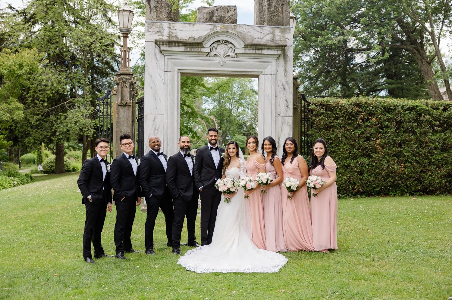 Wedding party posed under an ornate stone archway with bridesmaids in pink gowns and groomsmen in black tuxedos.
