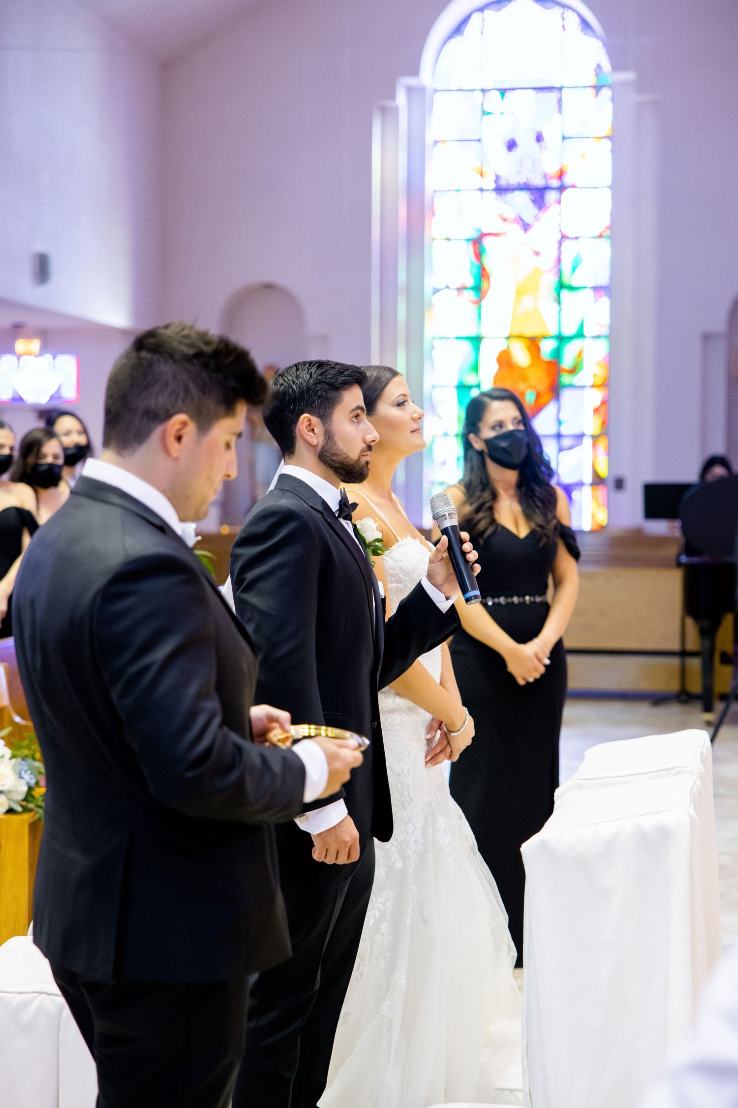 Wedding ceremony participants stand near stained glass window in church setting.