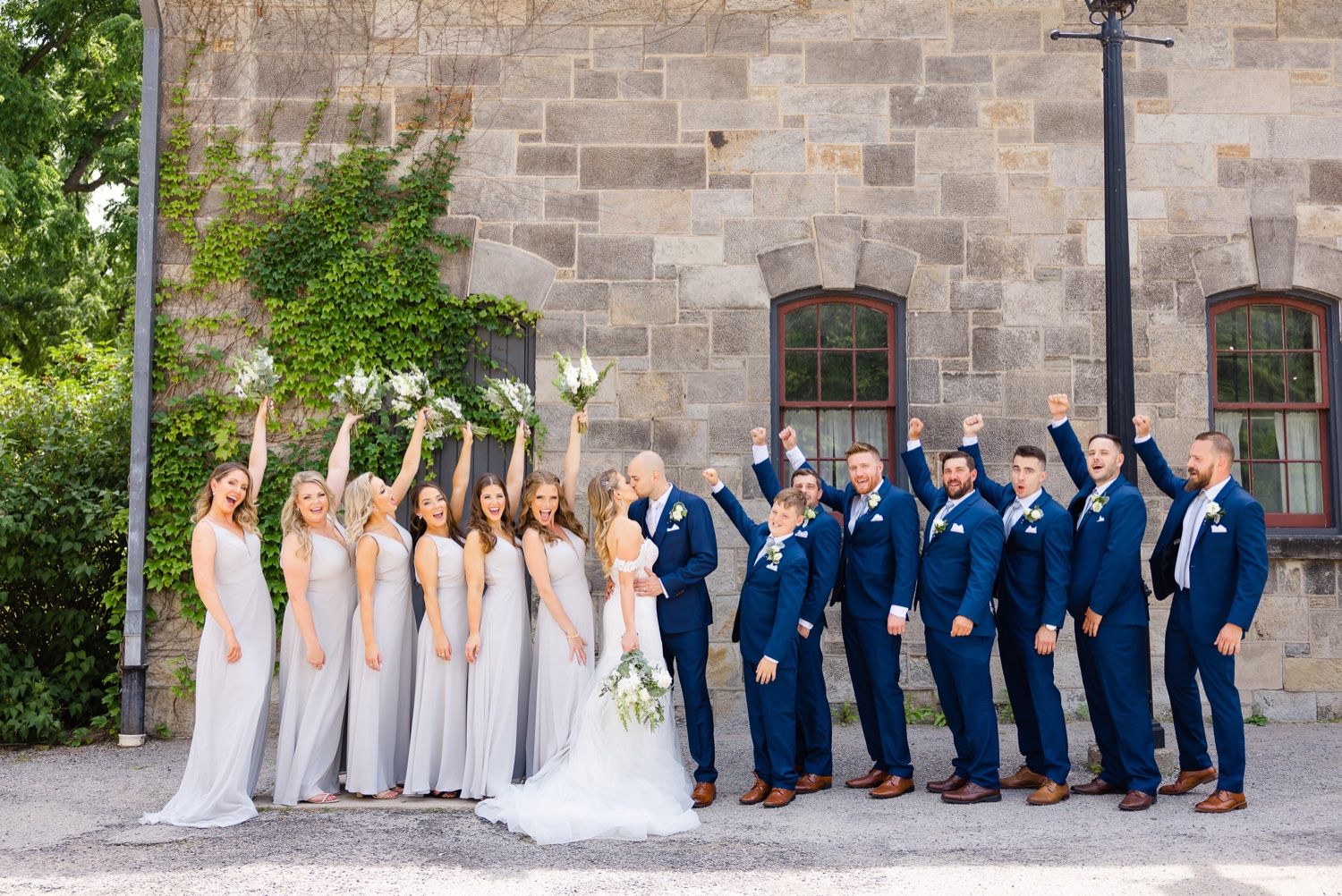 Wedding party in navy suits and white dresses celebrating with arms raised.
