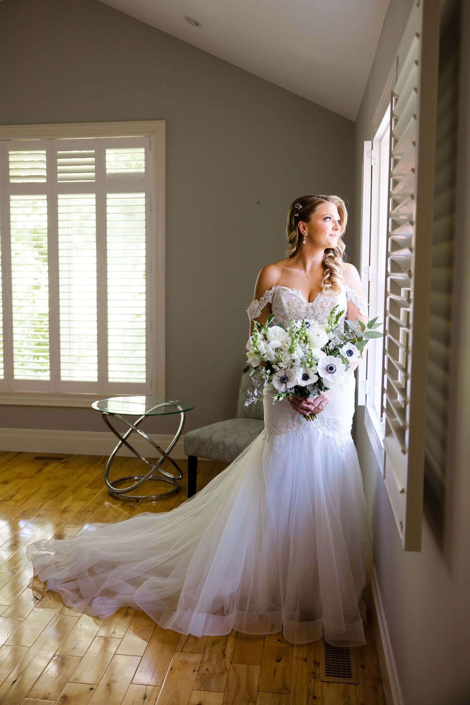 A bride holds a white bouquet while standing by a window in a flowing mermaid gown.