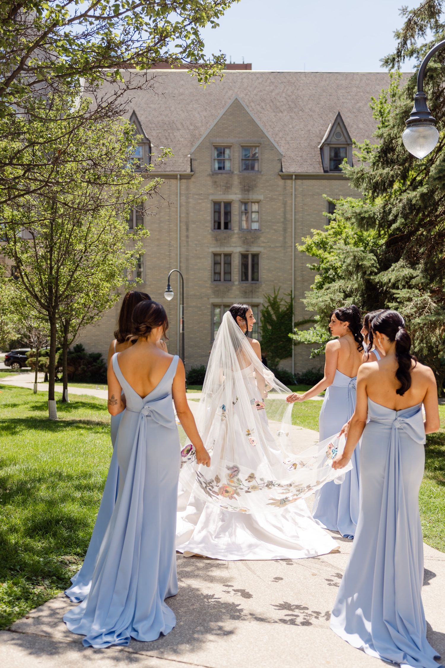 Bridesmaids in pale blue dresses hold bride's long veil on sunny garden path.