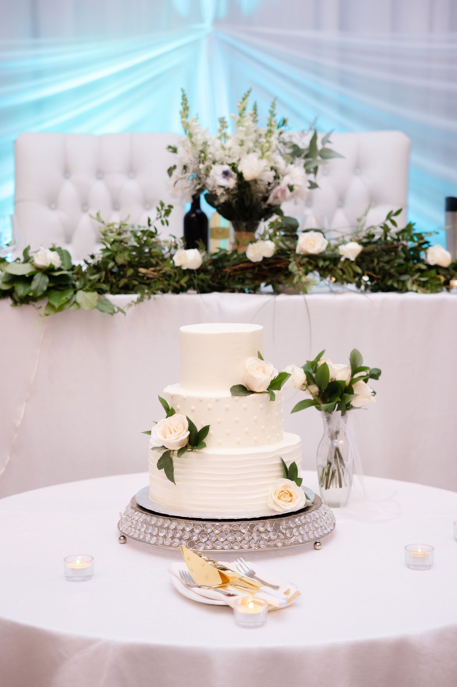 White wedding cake decorated with roses sits on silver stand against tufted backdrop with greenery.