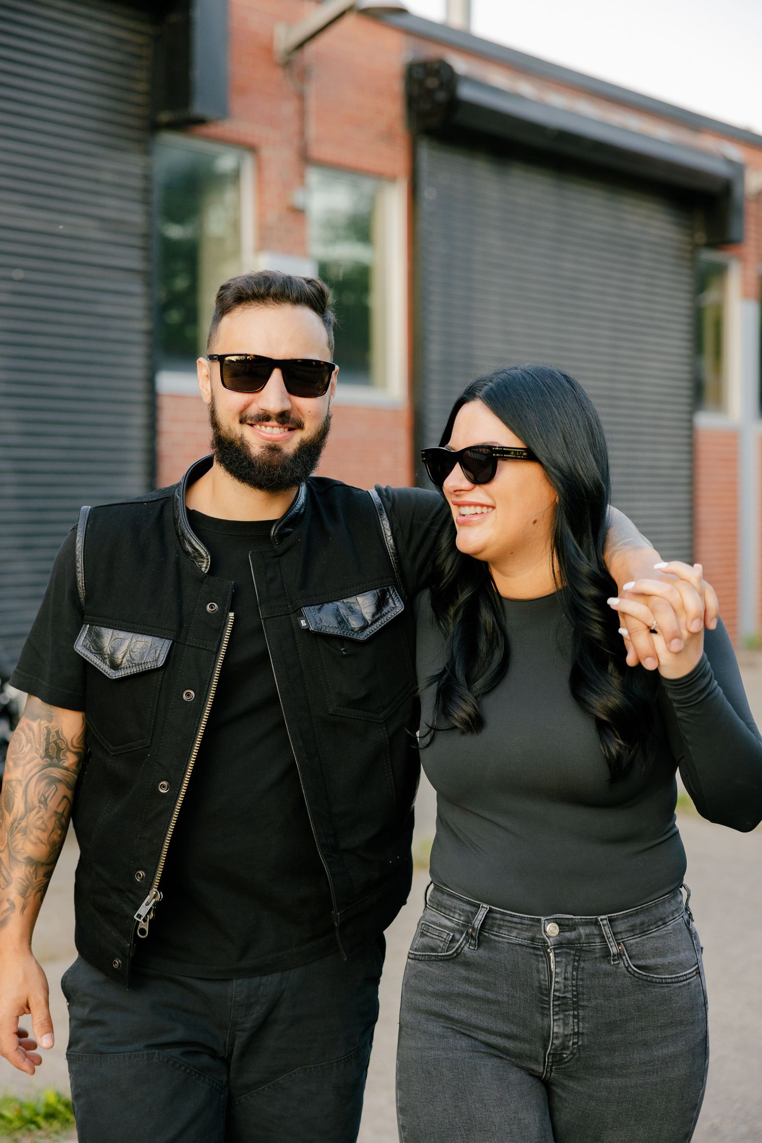 A couple wearing sunglasses and dark clothing smiling together against an industrial backdrop.