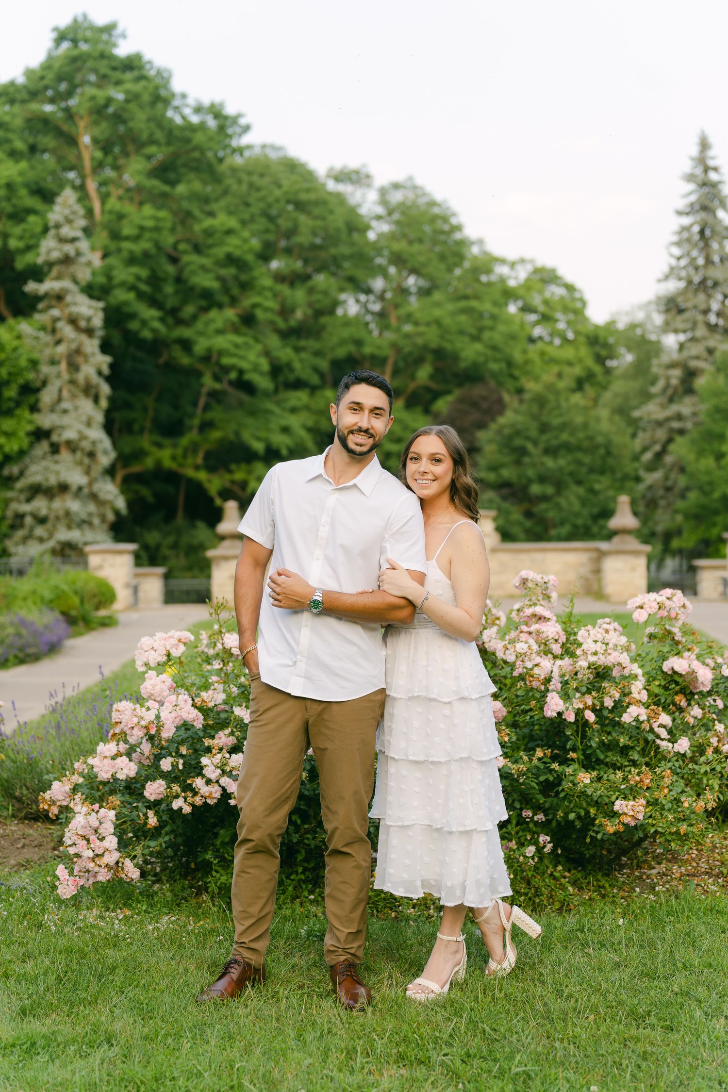 A couple poses together in a formal garden with stone pillars and blooming roses.