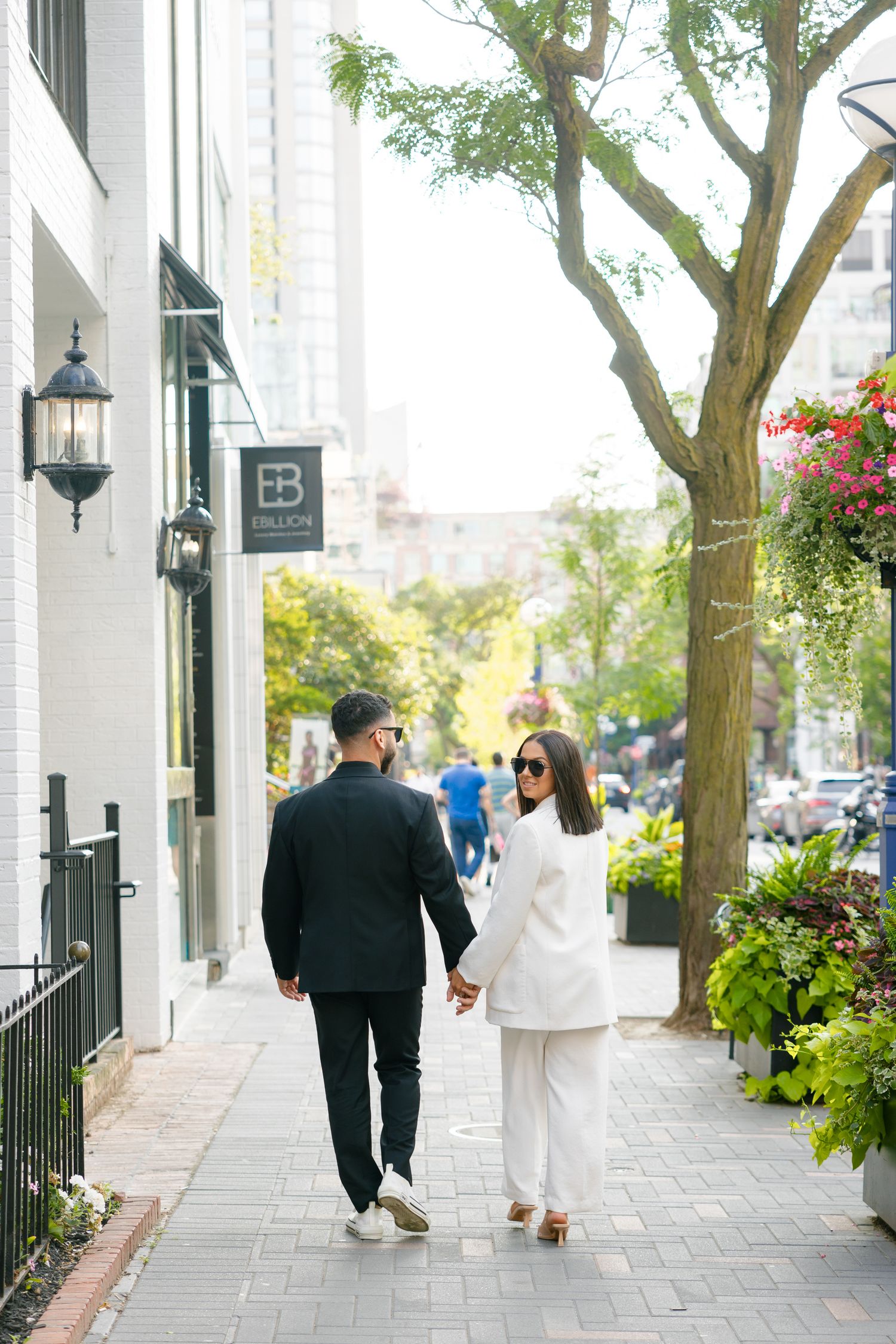 A couple walks along a tree-lined urban sidewalk with flowering planters and brick buildings.