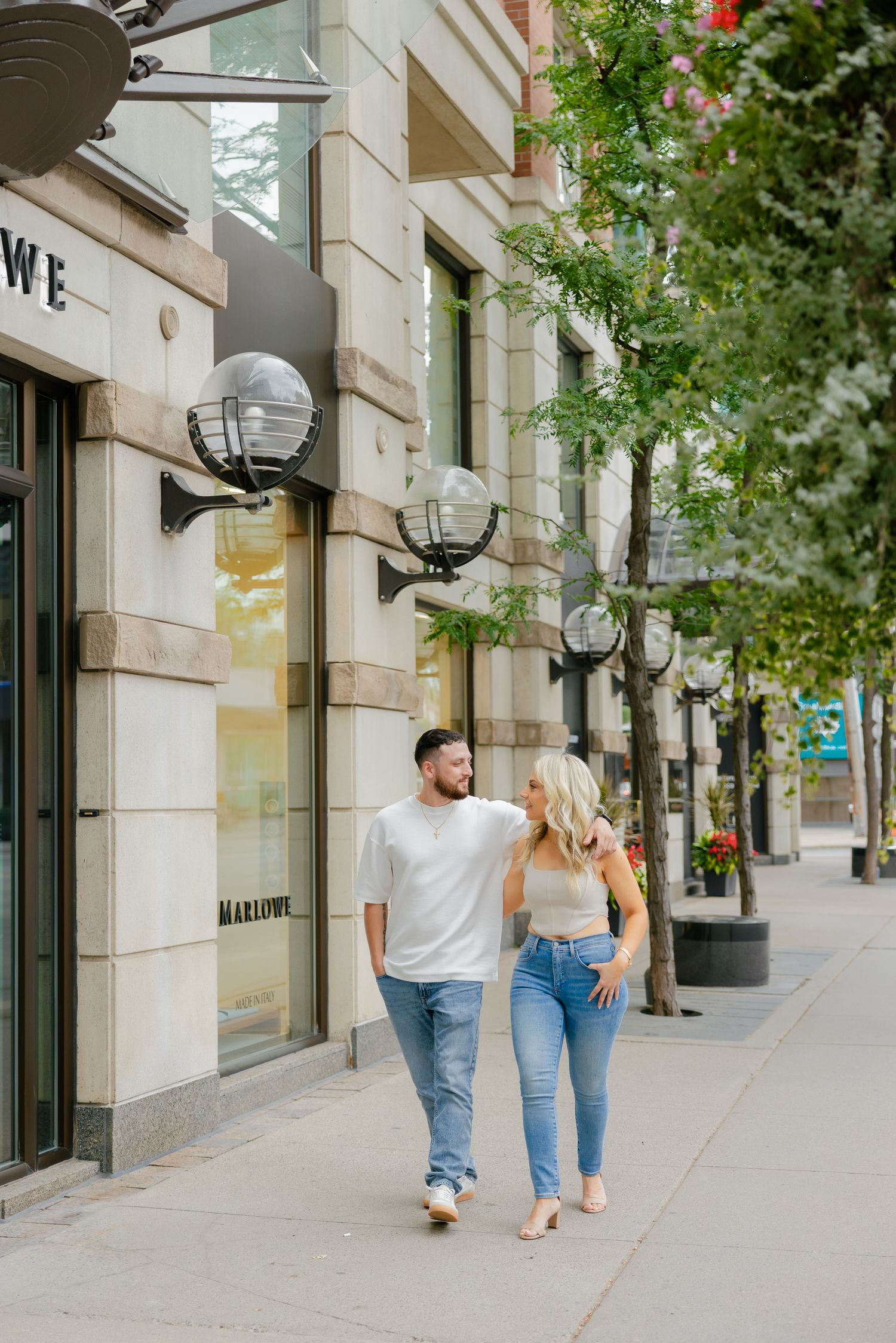 Couple walking together on urban sidewalk past storefront windows.