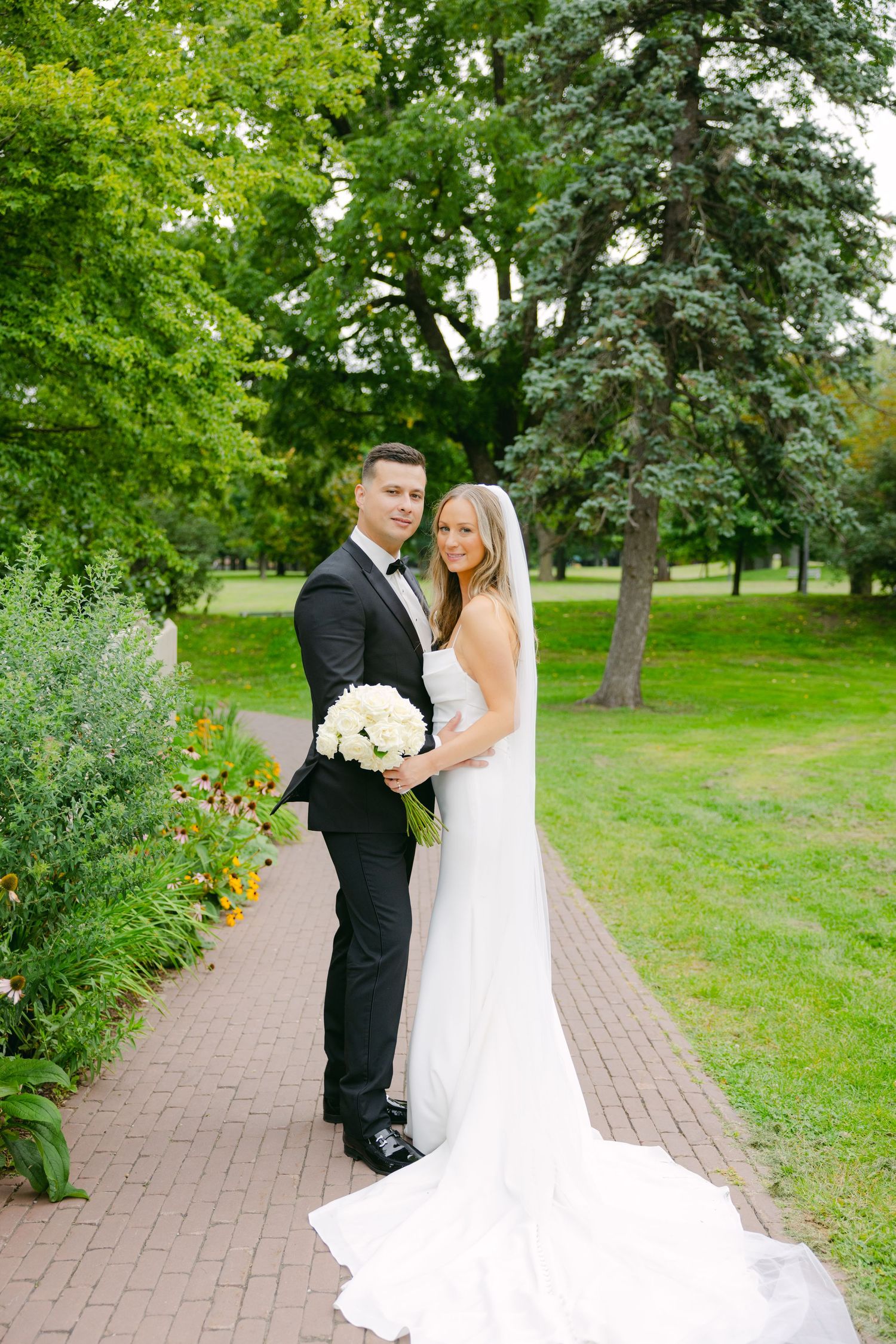 A couple in wedding attire poses on a garden path with white bouquet and flowing veil.