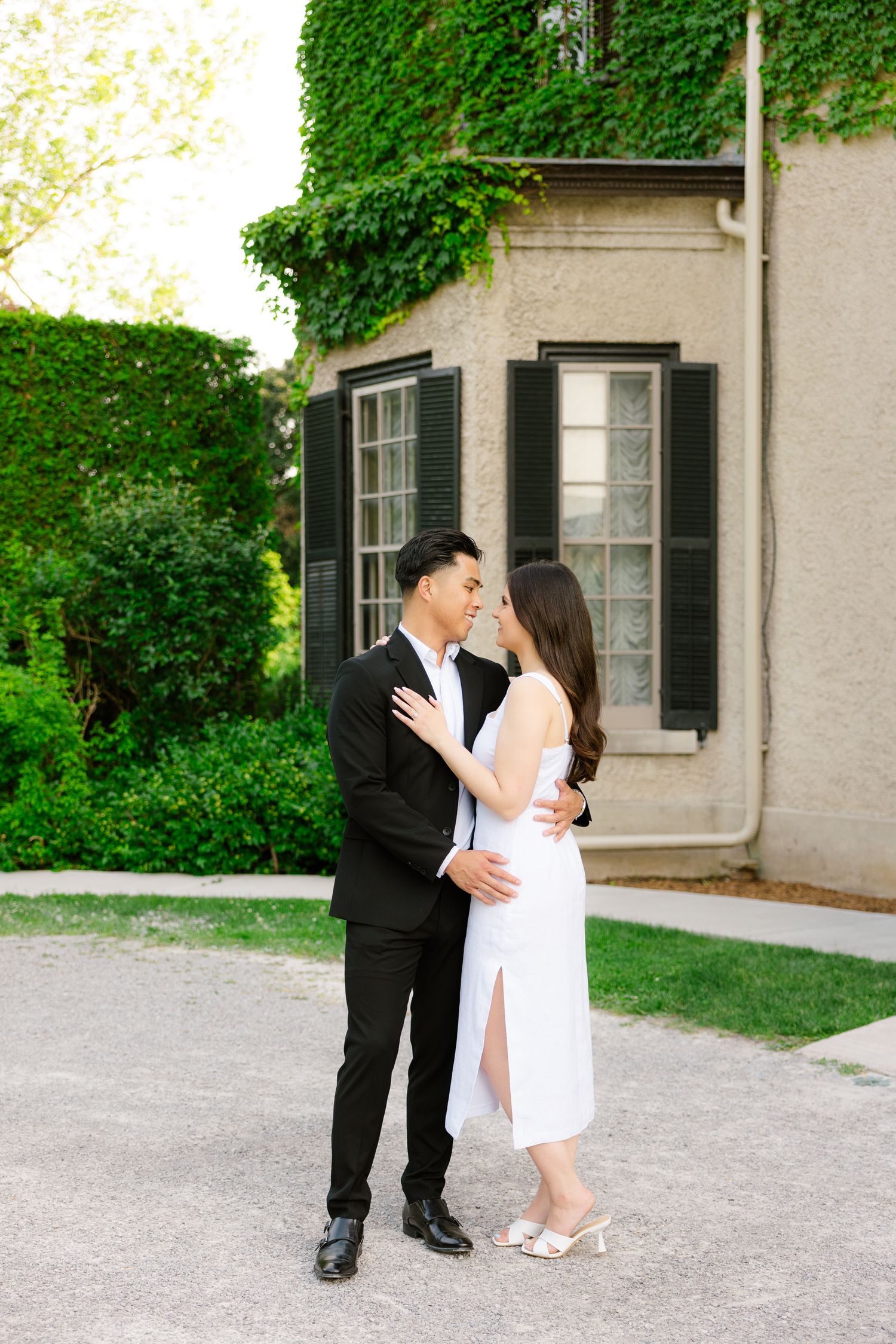 Elegant couple in formal wear posing against historic building with climbing ivy.