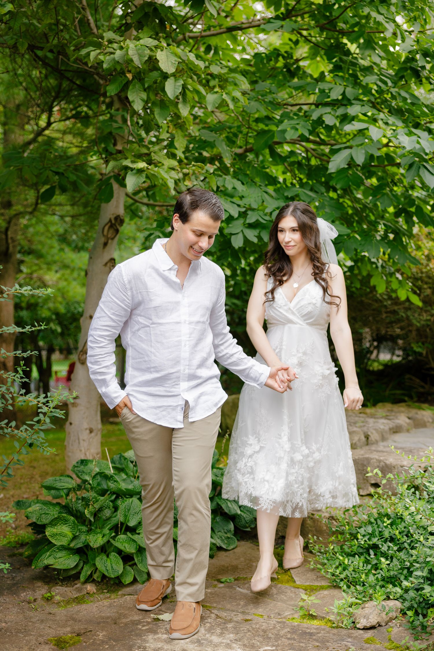 A couple walks together on a garden path surrounded by green foliage and trees.