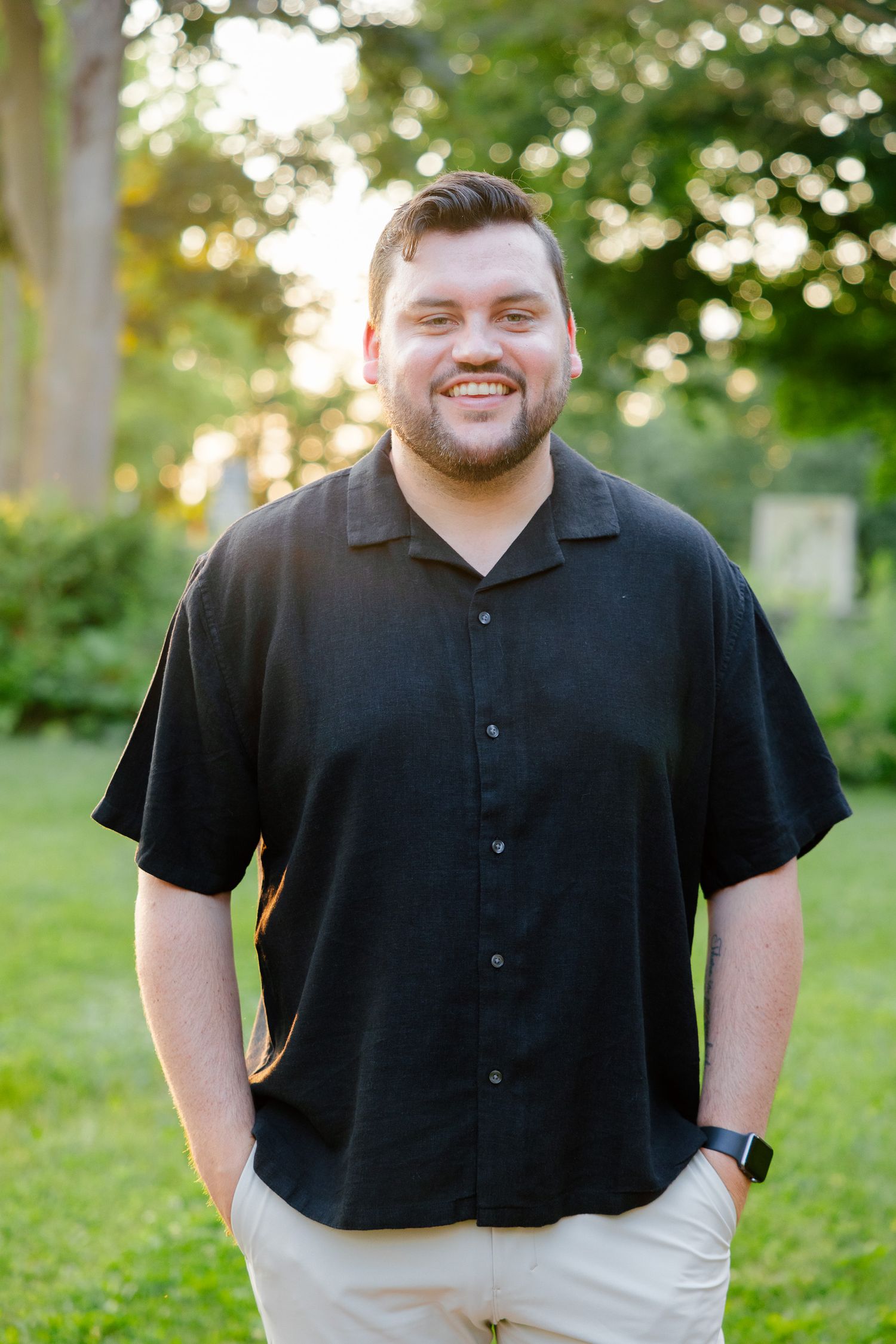 A person in casual attire smiles warmly while standing in a garden during sunset.