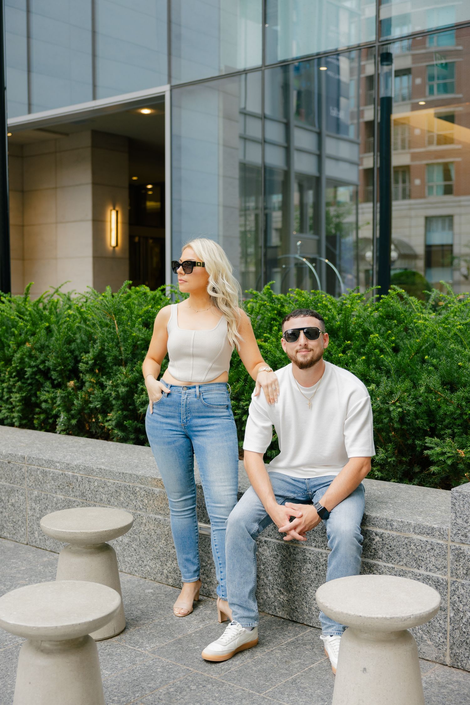 Trendy pair in casual attire pose near landscaped commercial building entrance.