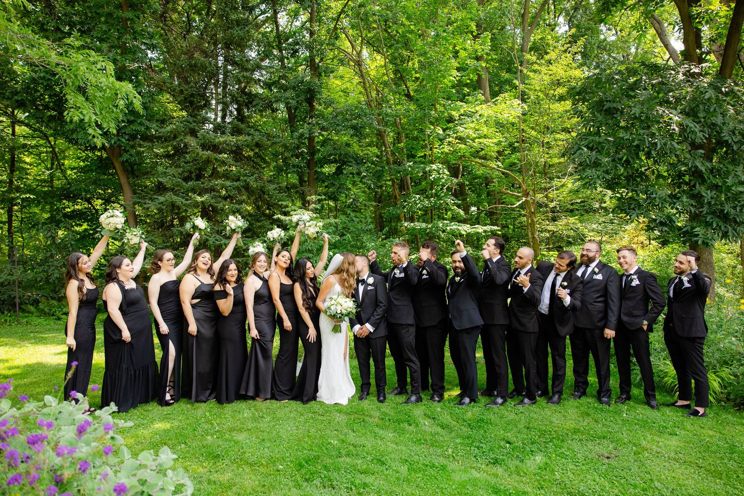 Wedding party celebrates on grass with arms raised while surrounded by lush summer greenery.