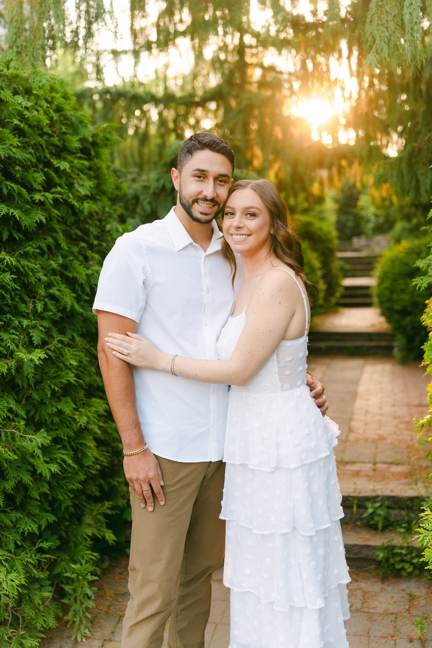 Happy couple poses together on a brick pathway surrounded by evergreen bushes at sunset.