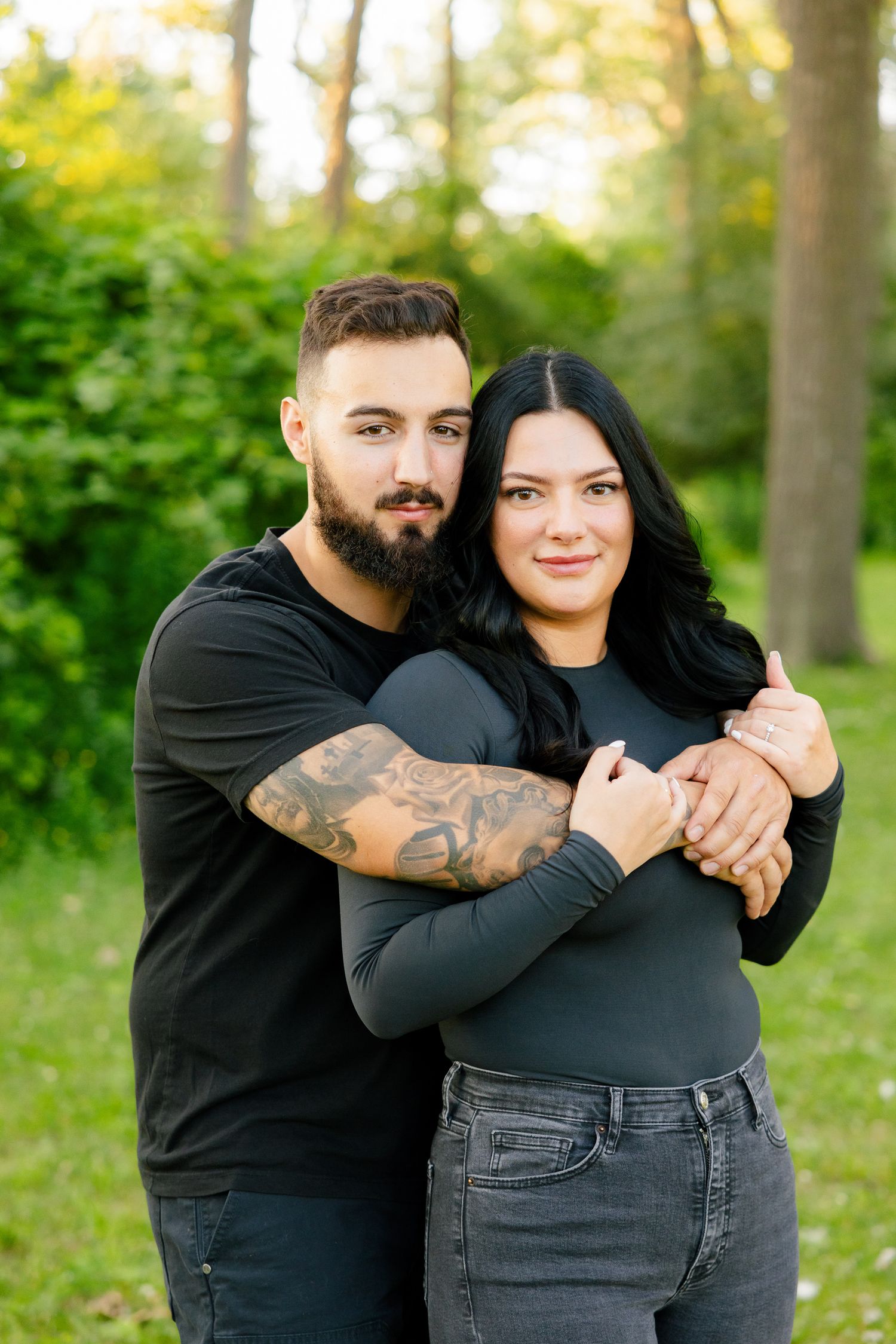 Outdoor portrait of couple in casual wear embracing against leafy green background.