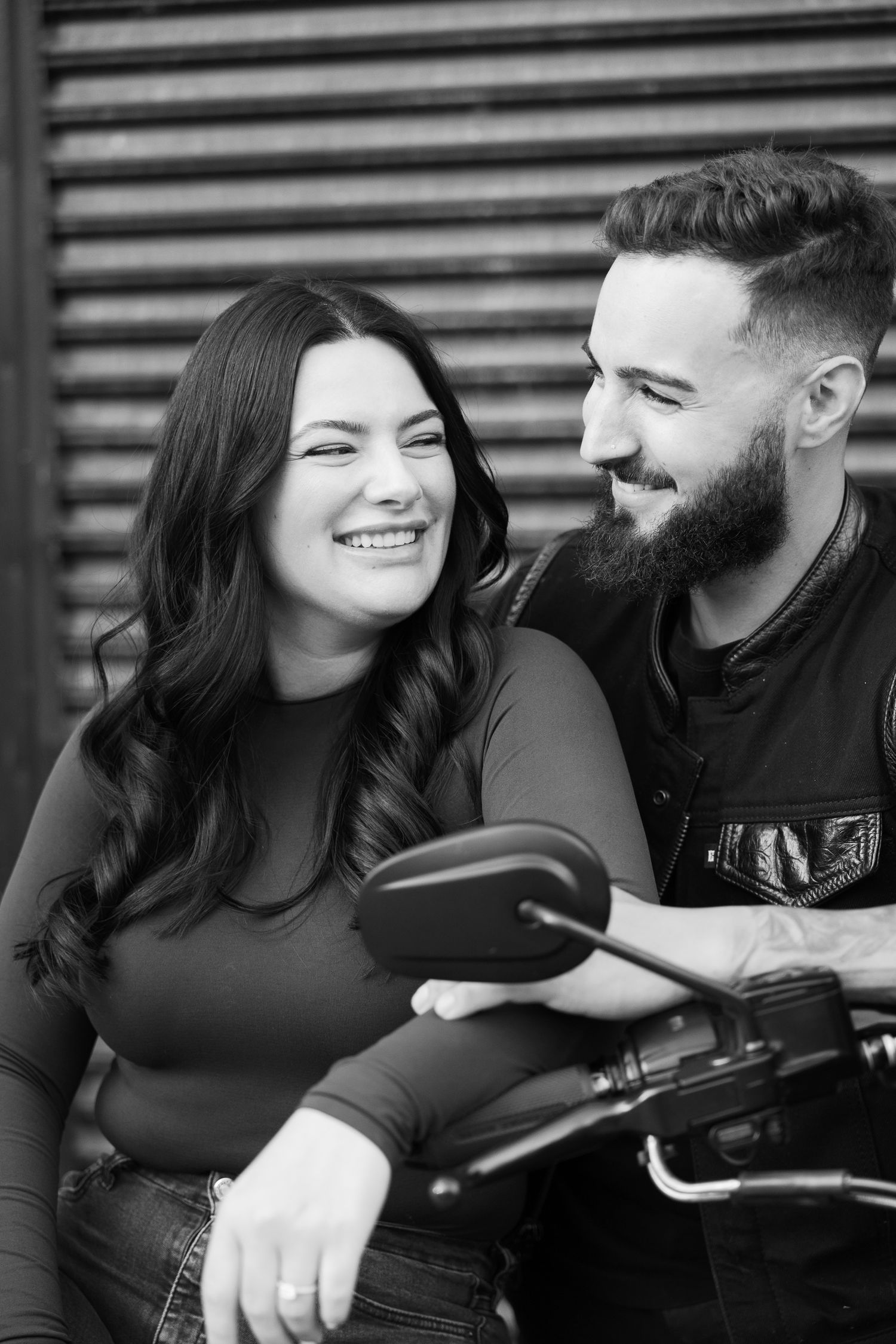 A joyful couple sharing a candid moment while leaning on a motorcycle against an industrial backdrop.