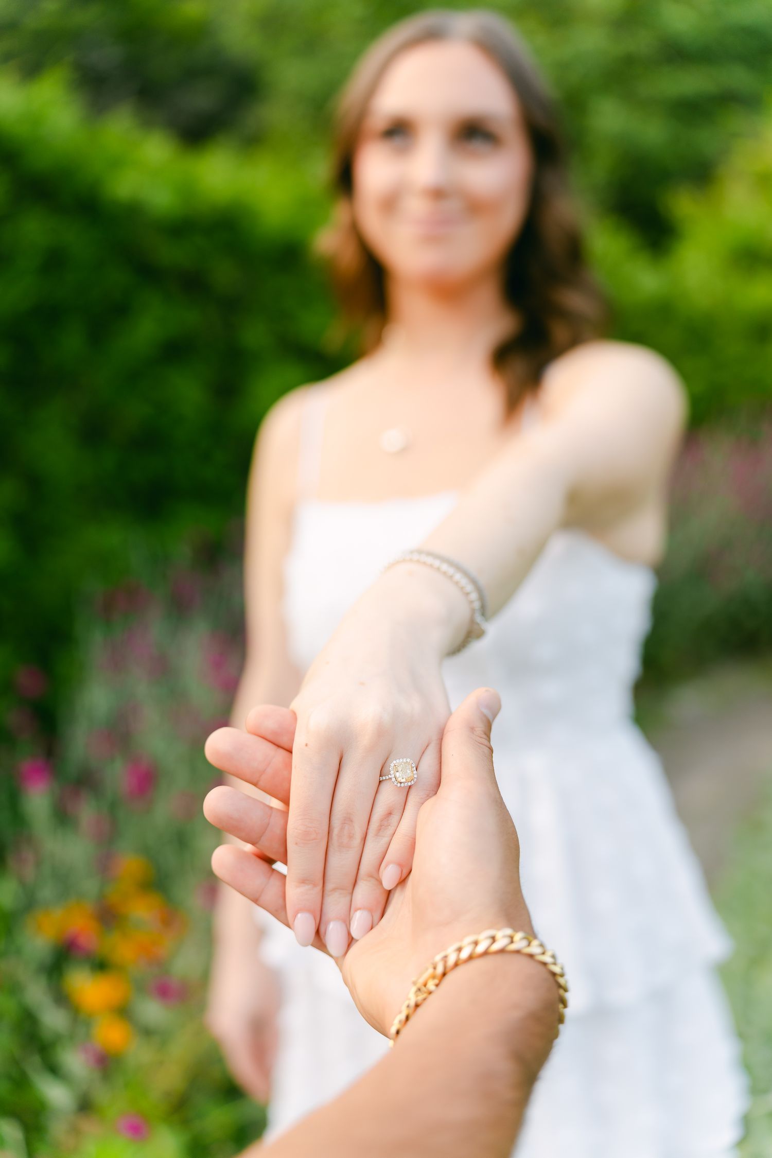 Close-up of hands holding engagement ring in garden setting with flowers.