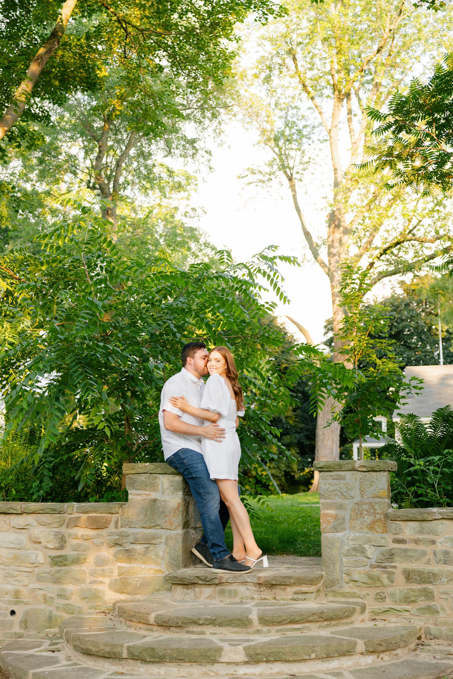 A couple embraces on curved stone steps in a garden setting with sunlit trees in the background.
