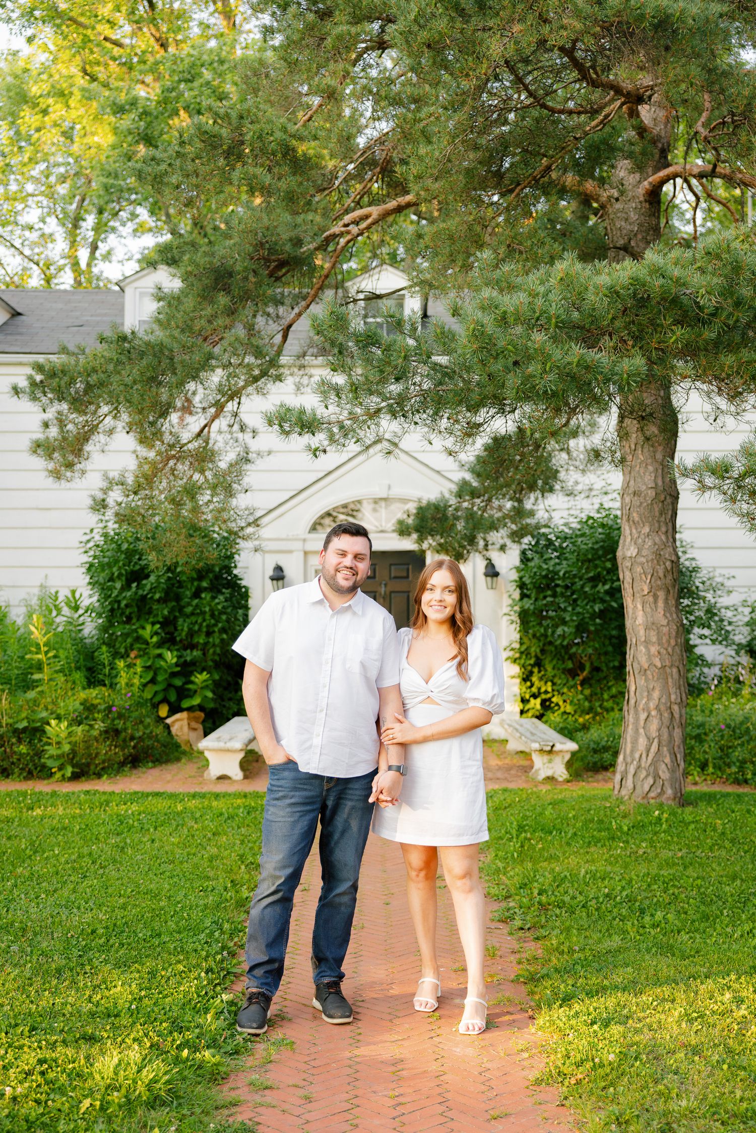 A couple poses on a brick walkway in front of a white building with large pine trees in the background.