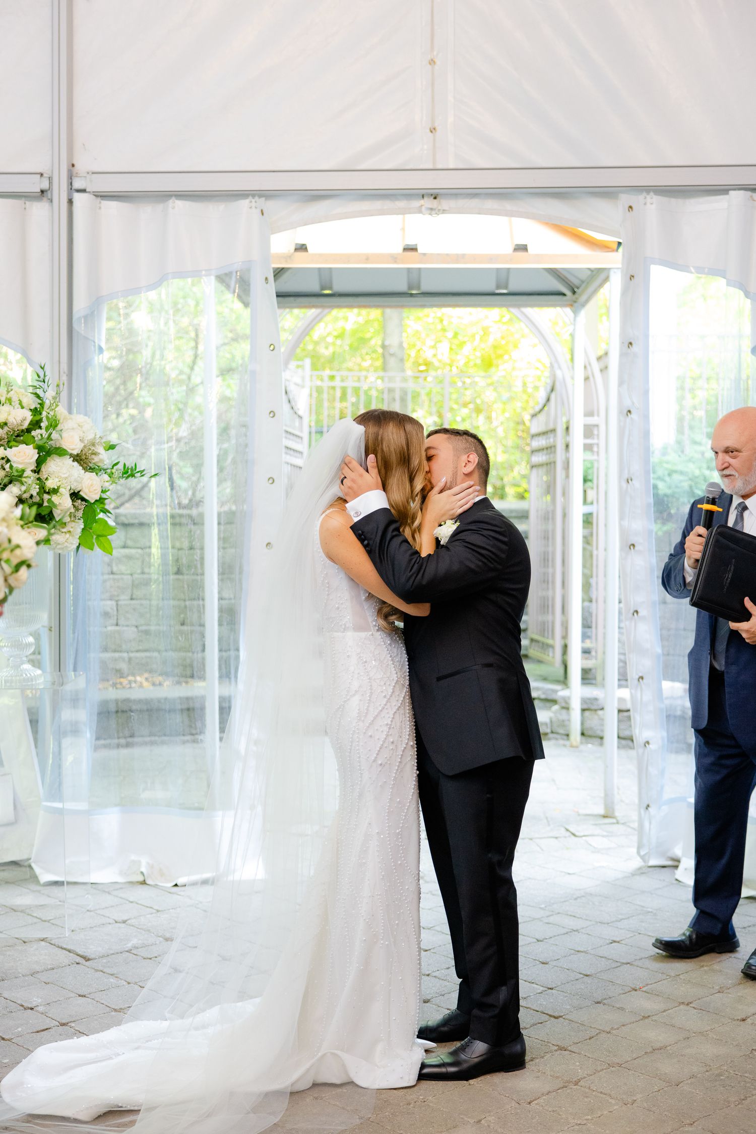 First kiss as newlyweds under white tent with officiant looking on.