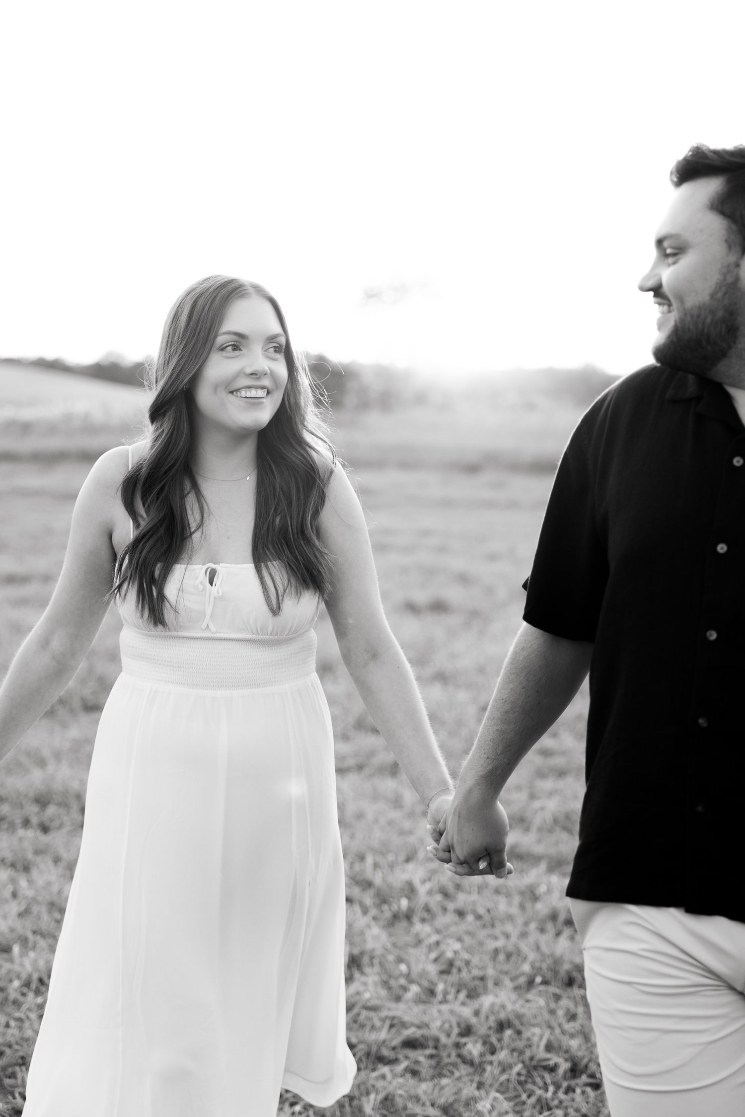 A black and white portrait of a couple sharing loving glances while holding hands in a field.