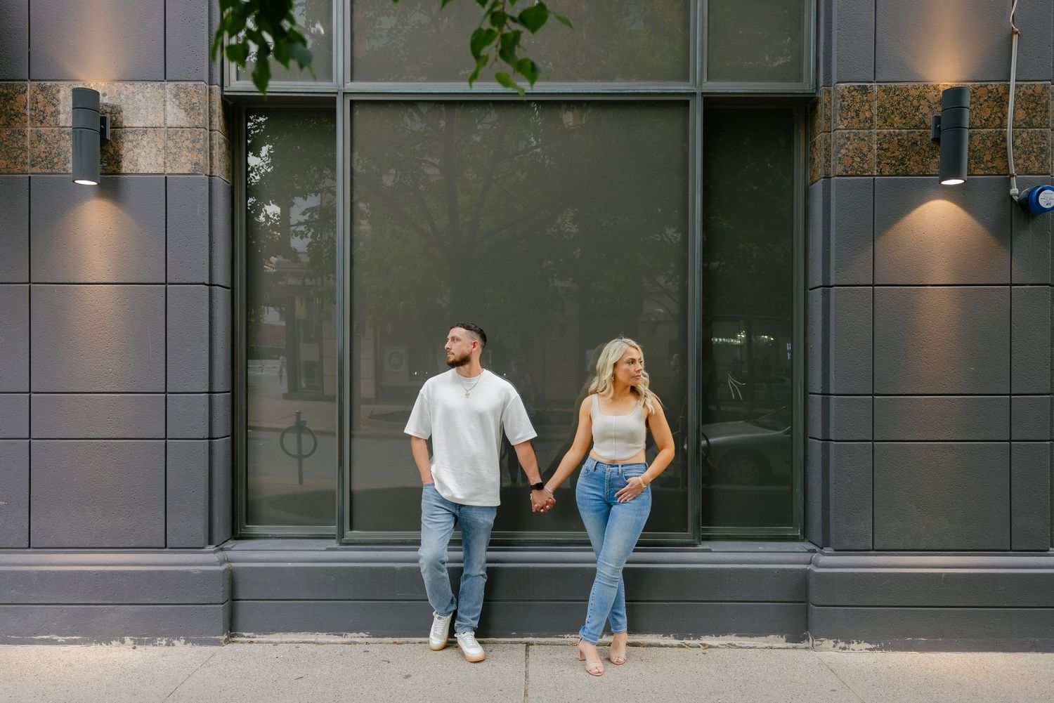 Modern couple posing casually against dark storefront window with architectural lighting.