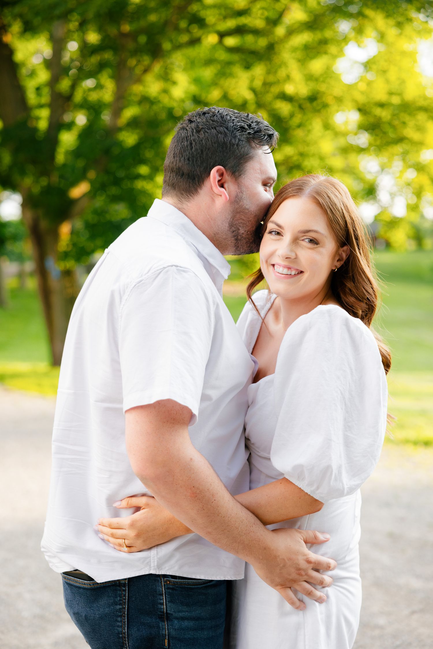 A couple shares an intimate embrace while standing on a path in a sunny park.