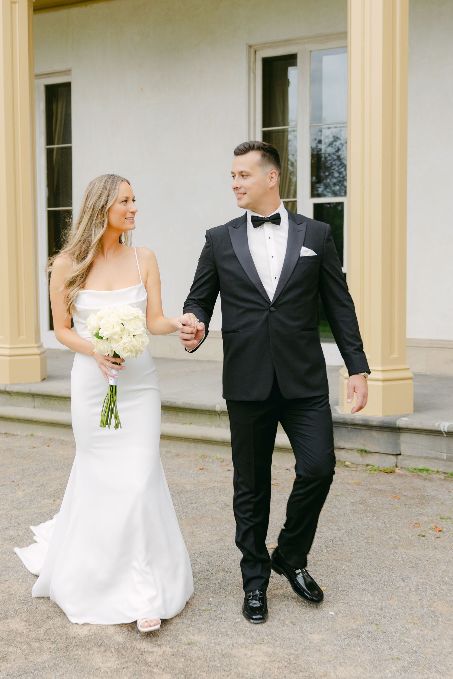 Bride with bouquet and groom walking together on classical porch with columns.