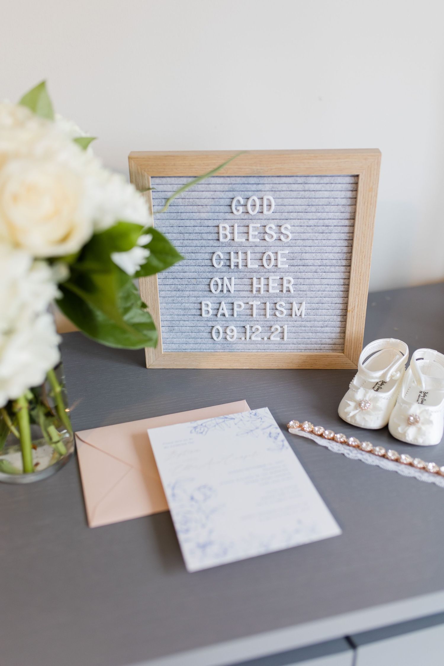Grey letterboard displaying baptism message with white roses and invitation on dark surface.