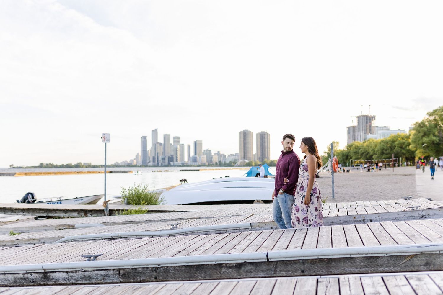 Couple standing on wooden boardwalk with city skyline visible in background.