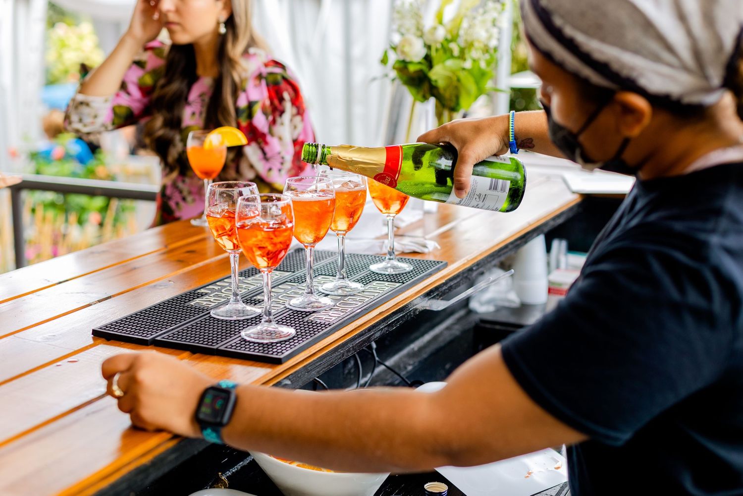 Colorful cocktails lined up on a wooden bar counter with bartender preparing drinks.