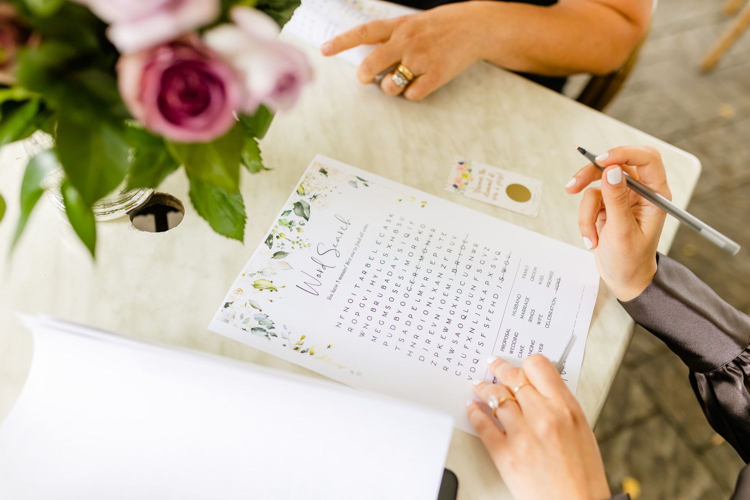 Hands filling out a document decorated with floral designs next to pink and purple flowers.