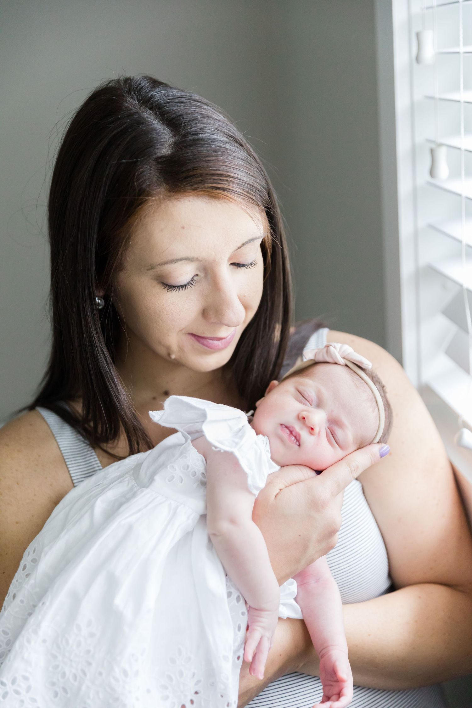 Tender moment between mother and newborn baby in white dress near window.