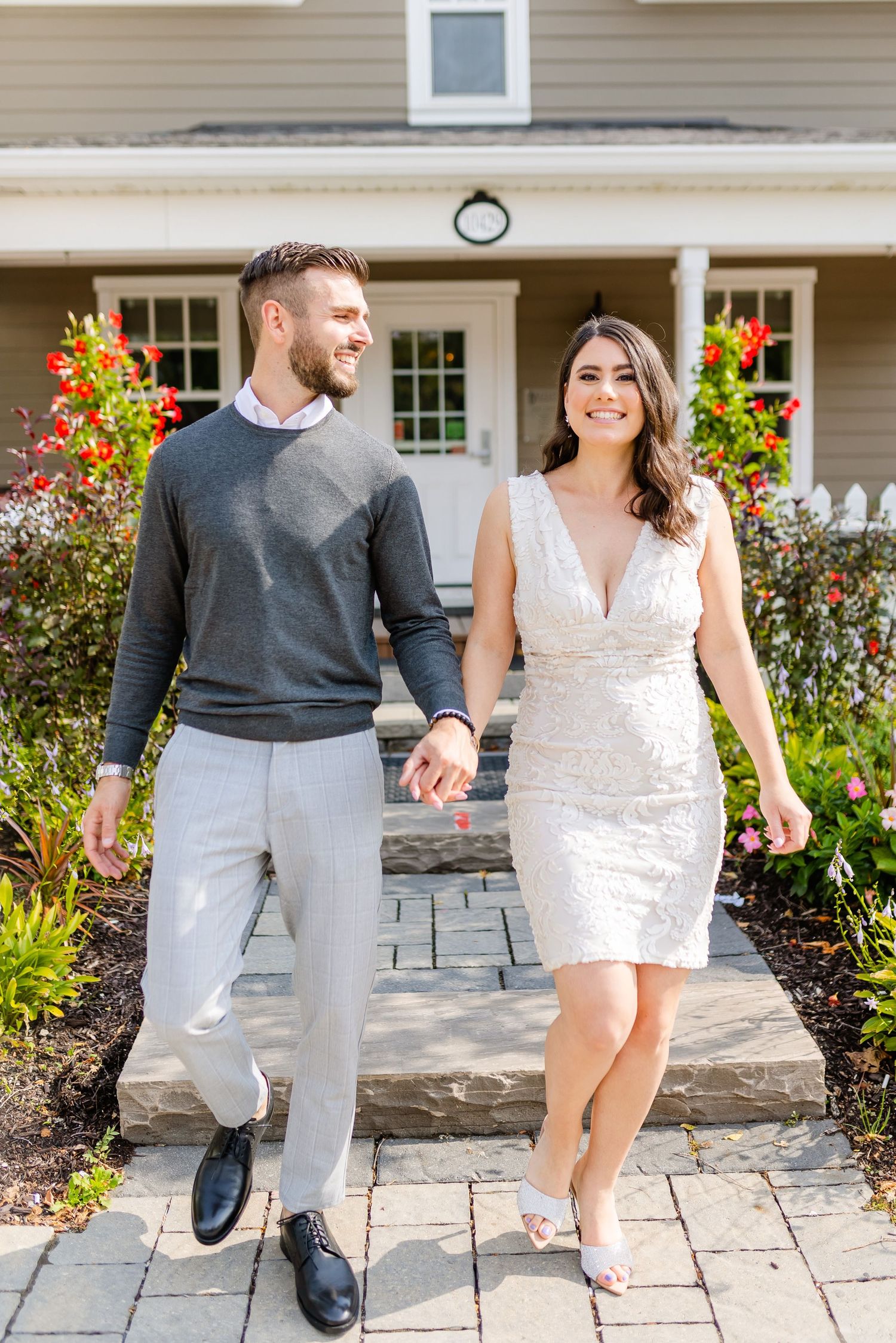 A couple holds hands while walking on a stone pathway in front of a house with colorful flower gardens.