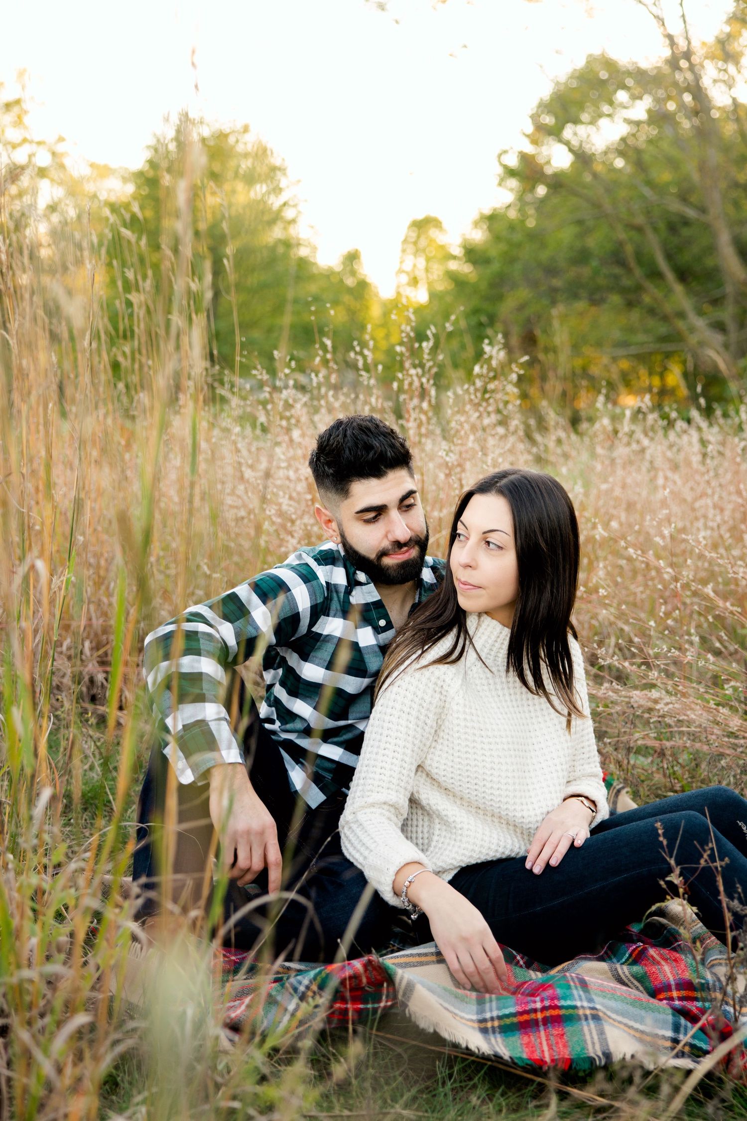Candid outdoor portrait of couple sitting on red plaid blanket among tall grass.