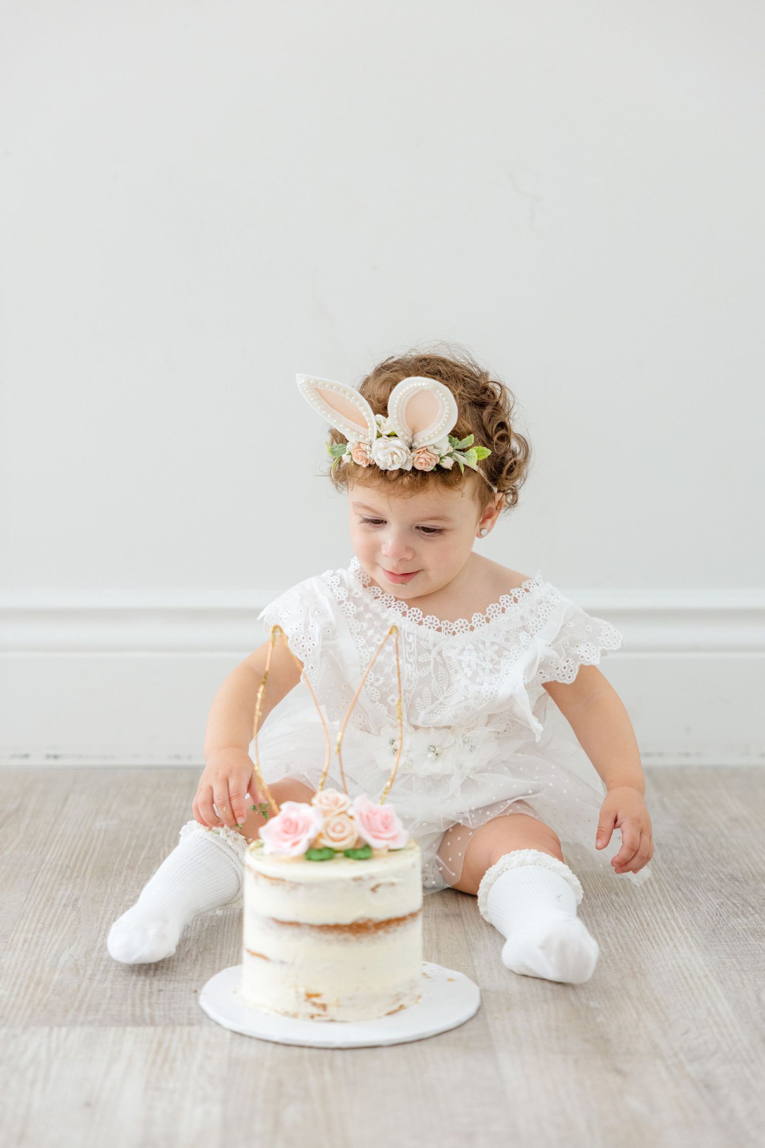 Sweet photoshoot scene with white lace dress and floral decorated cake on wooden floor.