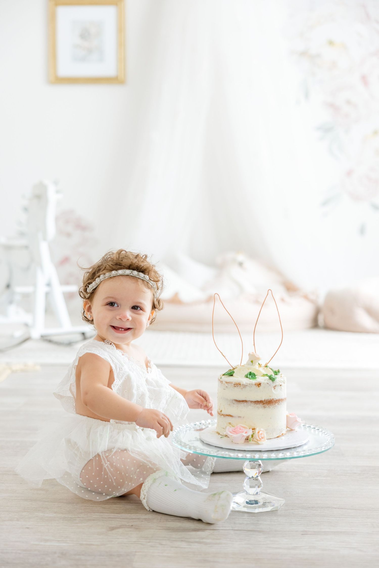 A child in white lace dress sits beside a delicate cake decorated with bunny ears and flowers.
