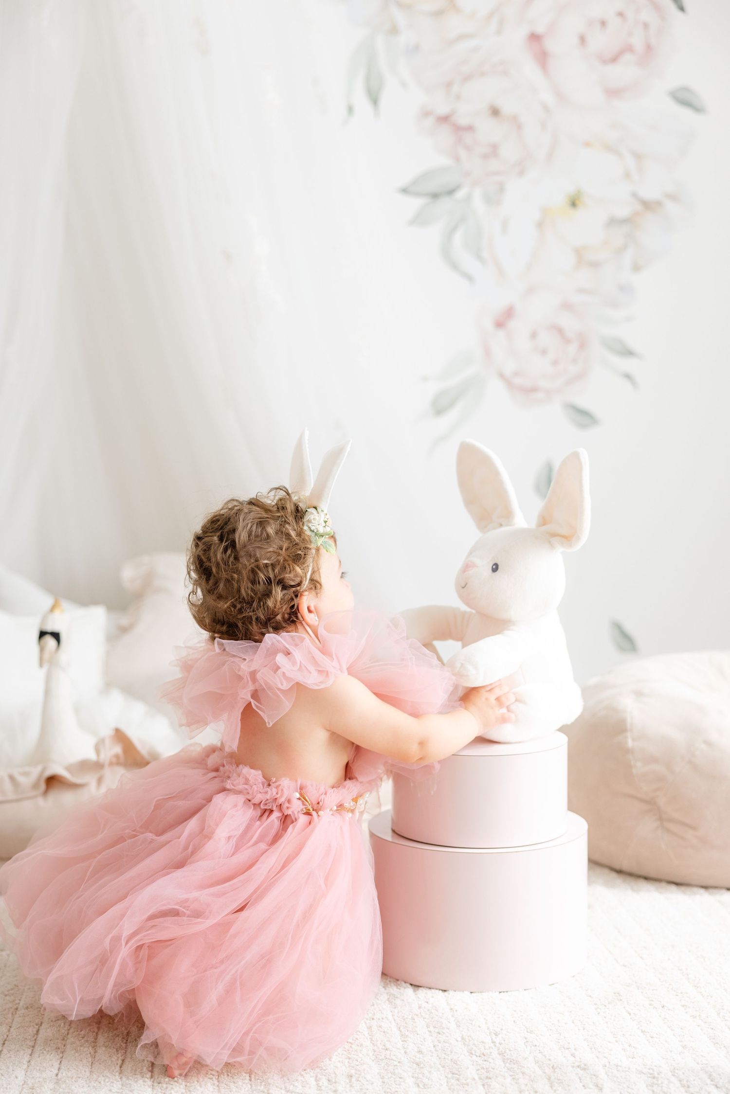 A toddler in a pink tulle dress reaches for a white bunny decoration in a soft, floral-decorated room.