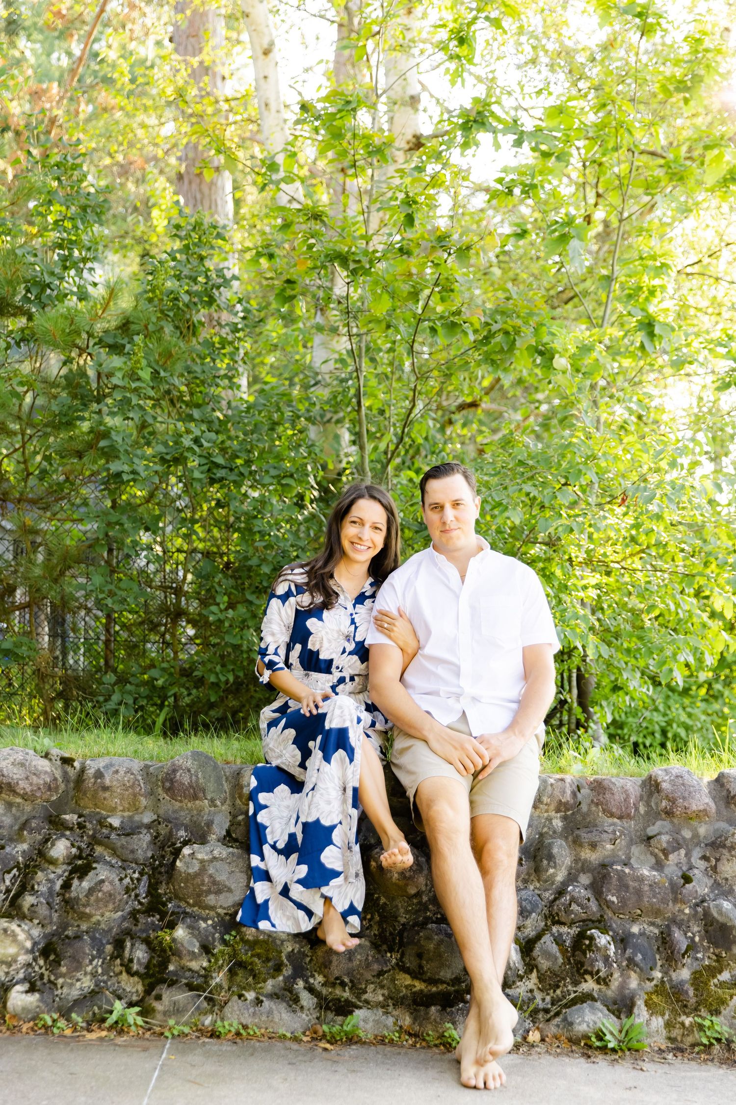 A couple poses against lush greenery while sitting on a stone wall in summer attire.