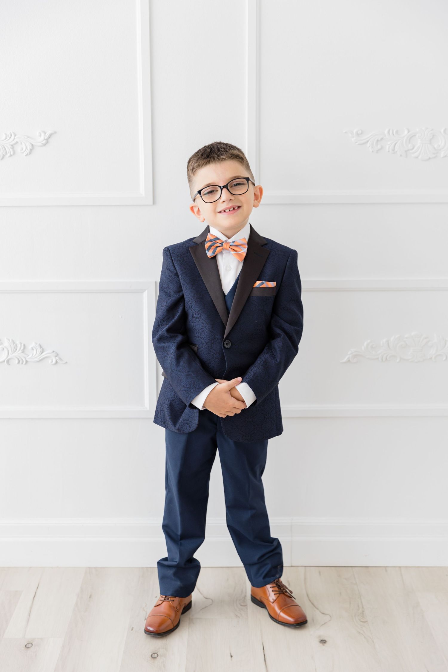 Full length portrait of youth in navy suit with orange bowtie and brown shoes against white wall.