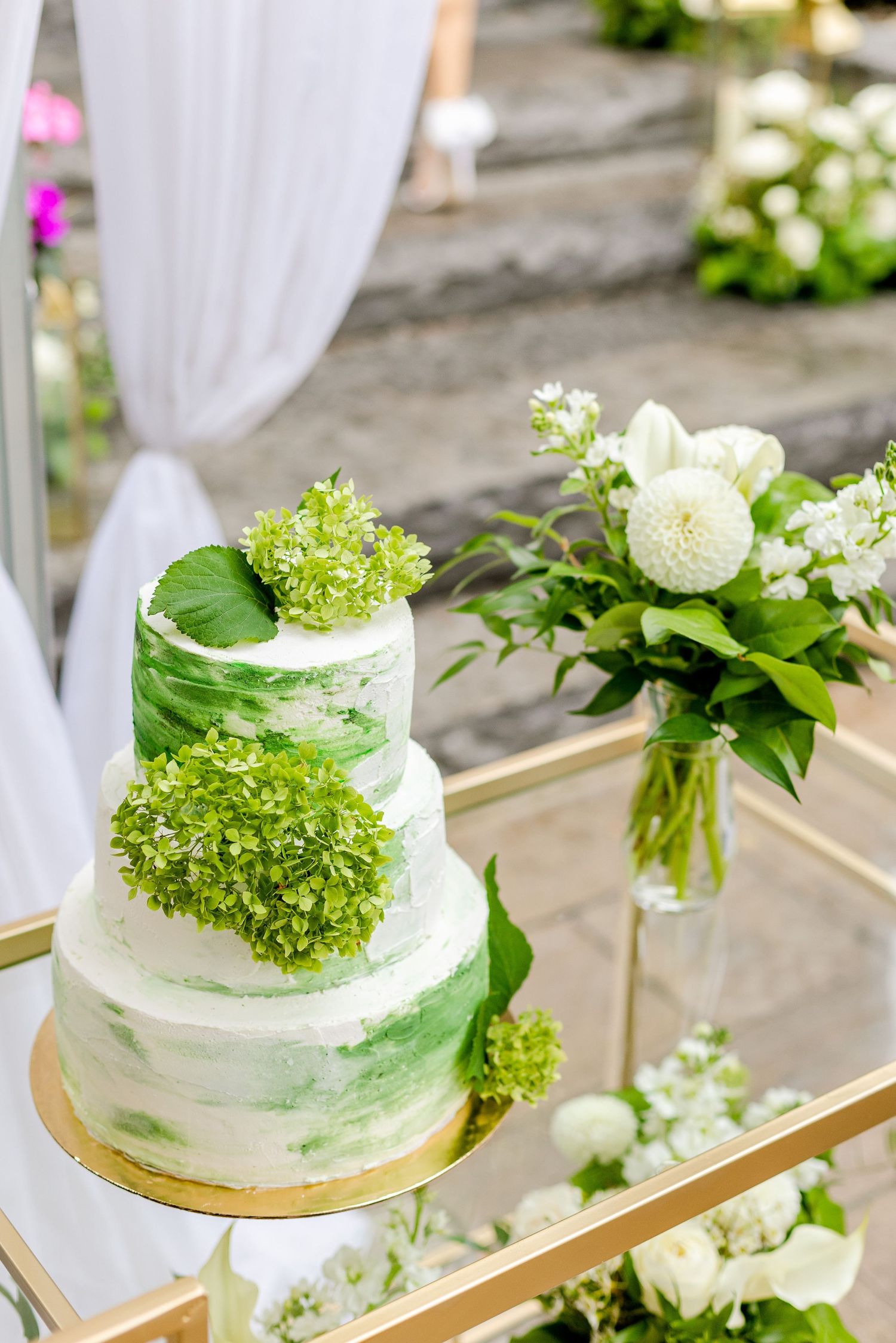 Modern white and green wedding cake displayed on gold mirror tray with fresh floral arrangements.